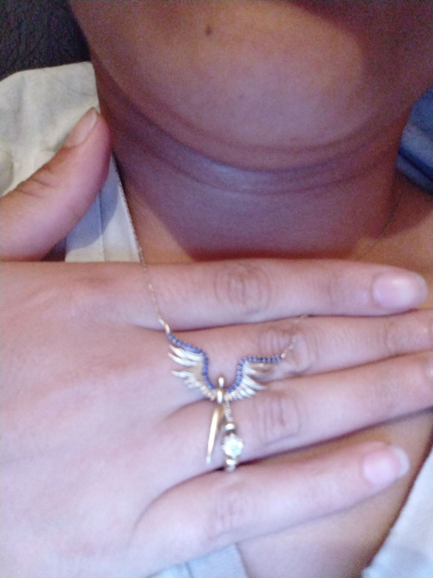 A close-up shot depicts a person's neck and upper chest area. Their hand is positioned across their chest and on the hand rests a necklace with a pendant resembling a winged figure. The wings of the figure are adorned with blue stones, and the pendant features a white stone or pearl at the bottom. The background is blurred, showing the collar of a light-colored shirt.