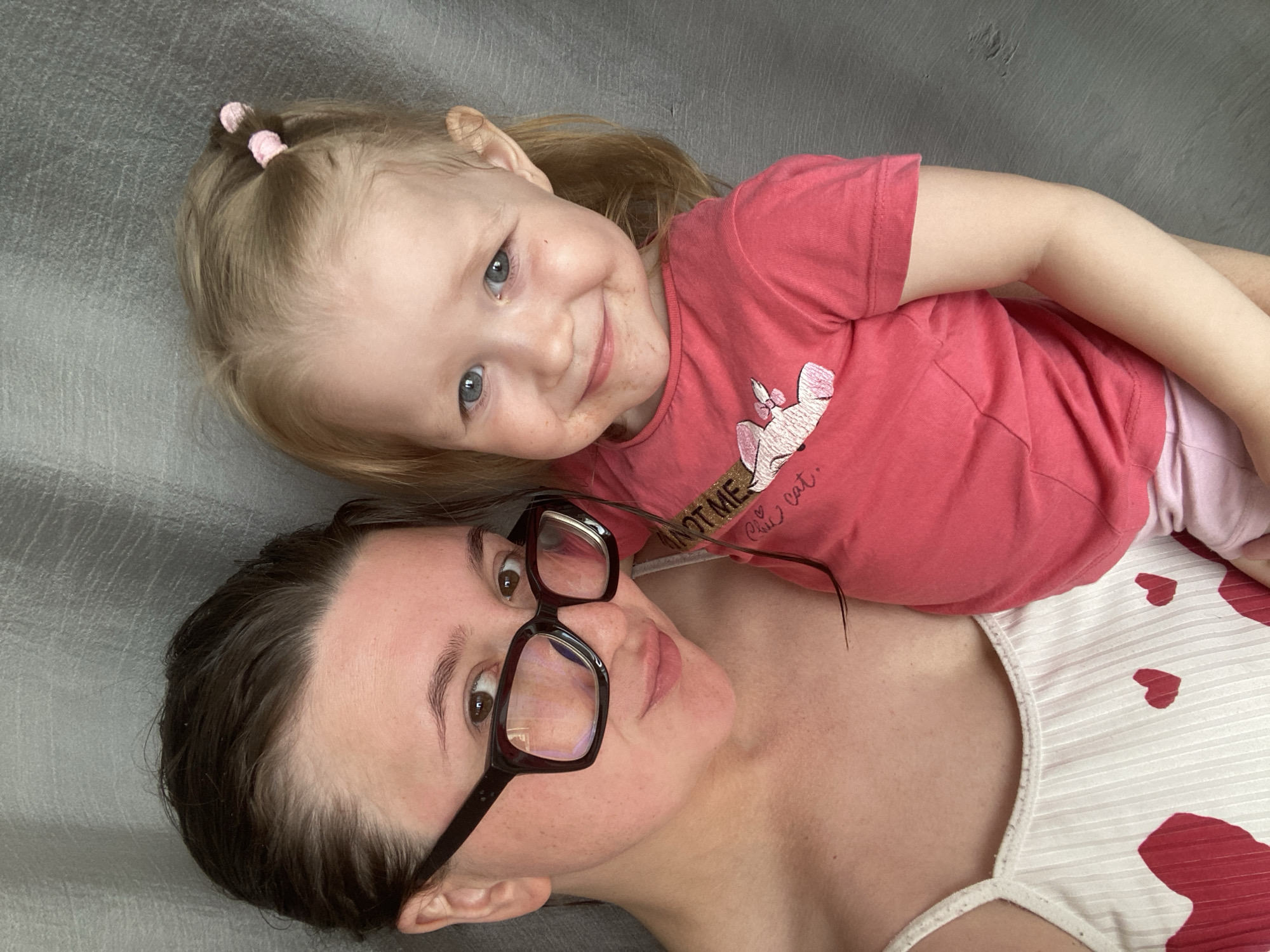 The media file is a close-up, overhead shot of two females, likely a mother and her daughter, lying down together. The older female, on the bottom, is wearing black-rimmed glasses and a white top with red heart patterns. She has dark brown hair and is looking towards the camera with a neutral expression. The younger female, on top, has blonde hair tied in two small ponytails with pink hair ties. She is wearing a pink t-shirt with a graphic print and is smiling directly at the camera, her face appearing slightly smudged. The background appears to be a textured gray surface, possibly a couch or bed. The lighting suggests it's daytime, and the overall mood is intimate and affectionate. The text "NOT ME" and "Cute cat." are visible on the younger female's t-shirt. The location context is Chaikovsky, Russia.