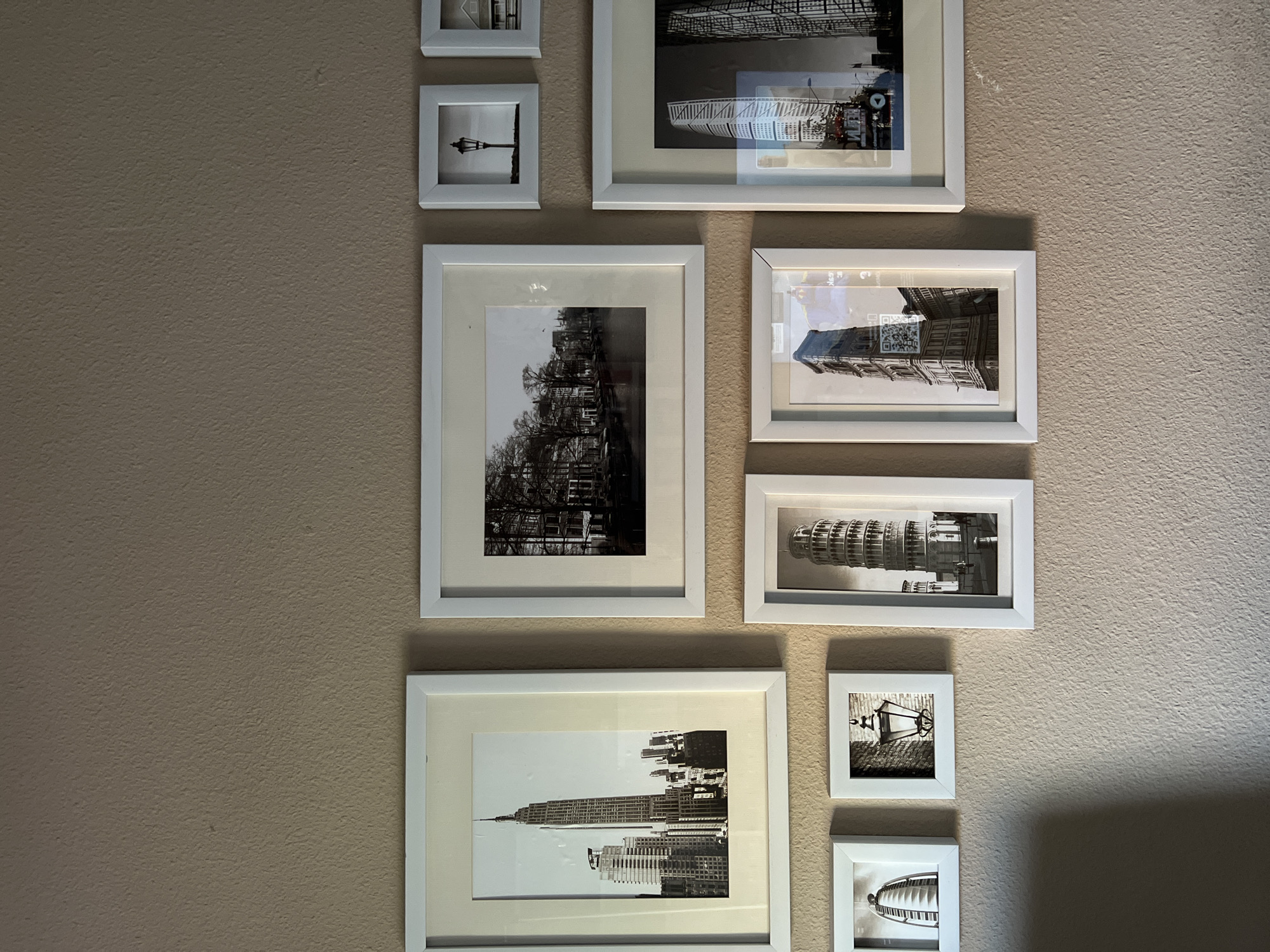 A collection of framed black and white photographs hangs on a beige wall. The photographs are of buildings and architectural landmarks. The frames are all white, and arranged vertically. The images include the Empire State Building, the Leaning Tower of Pisa, and architectural scenes of buildings in Amsterdam, Netherlands, and Venice, Italy.