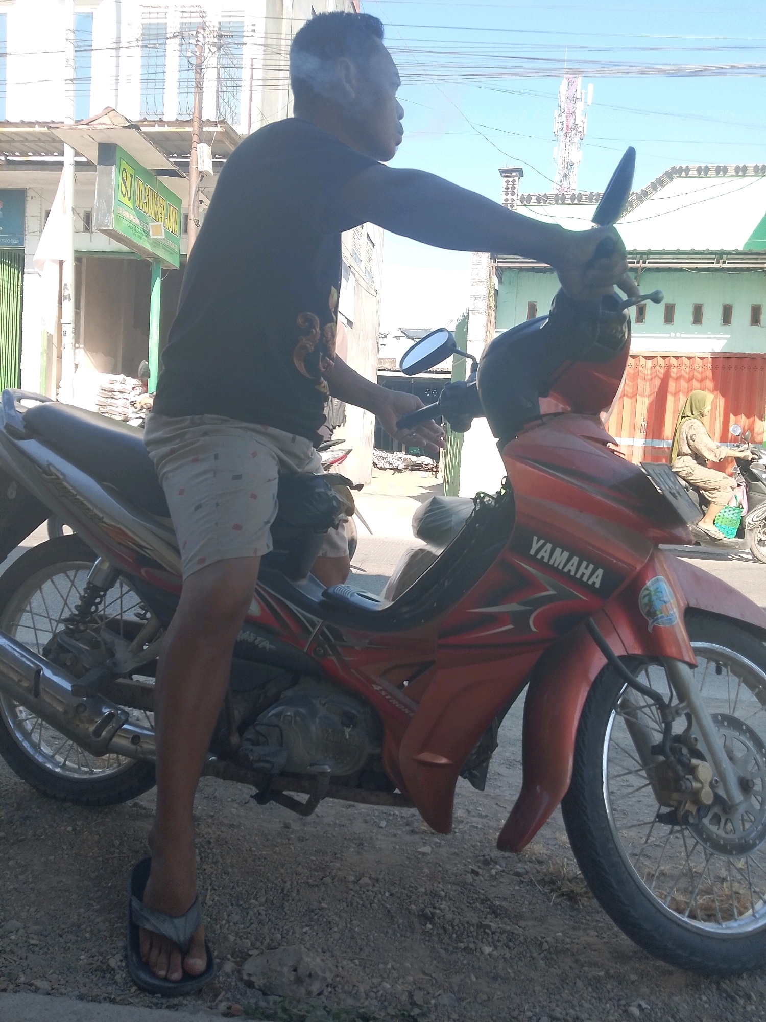 A man is sitting on a red and black Yamaha motorcycle in Jampet, Indonesia. He is wearing a black t-shirt, patterned shorts, and flip-flops. He is smoking a cigarette and appears to be looking to the right. The motorcycle is parked on a dirt road, with buildings and a cell tower visible in the background. Another person is riding a motorcycle in the background. The weather is clear and sunny.