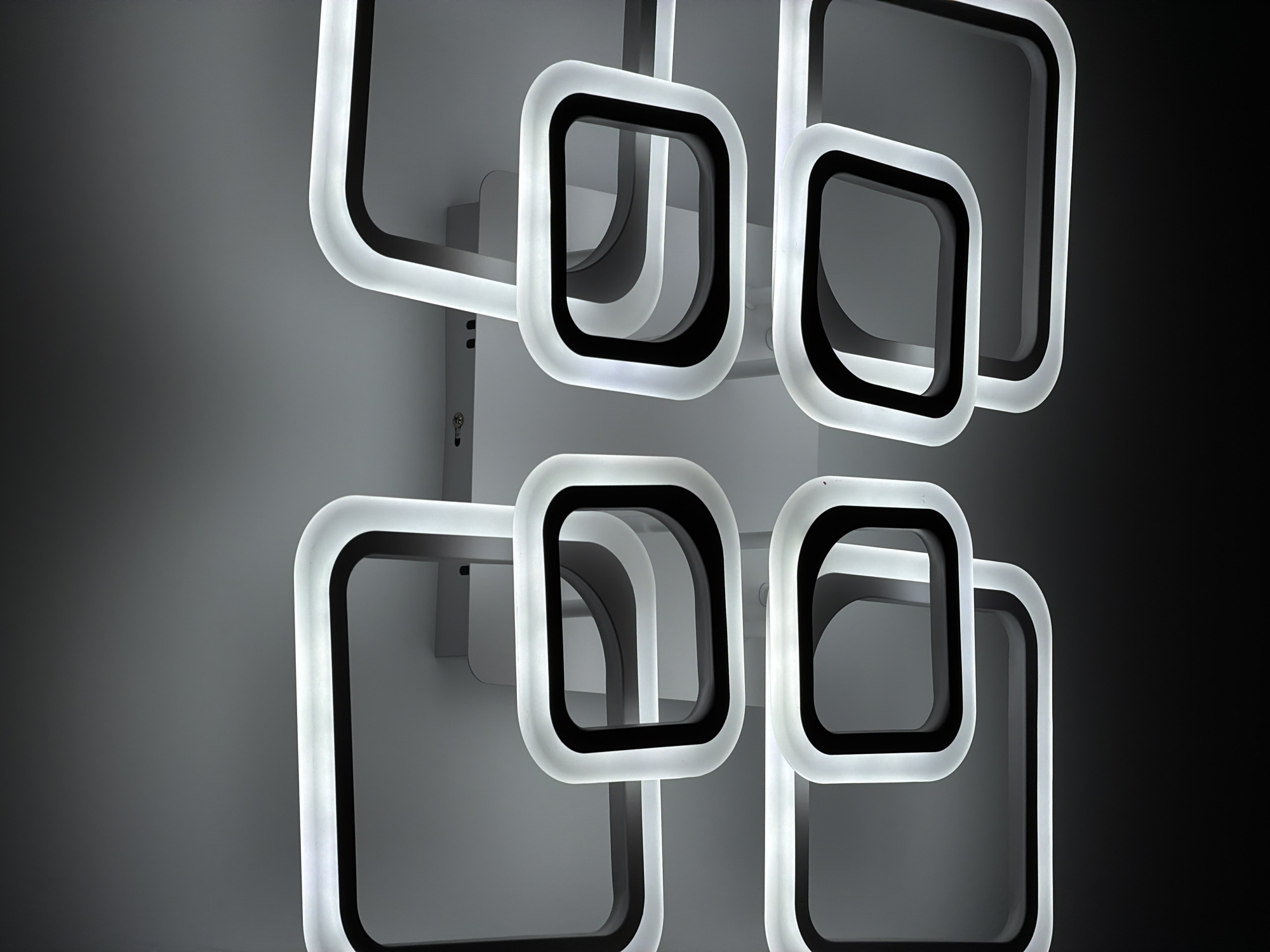 The image presents a close-up view of modern LED light fixtures, showcasing their geometric design and illumination. The primary subjects are multiple square-like elements with smoothly rounded corners, arranged in an overlapping, dynamic composition. Each element features a prominent black inner frame that contrasts sharply with an outer, translucent white border, which is brightly illuminated.
These individual light units are mounted on a light gray wall or vertical surface, appearing to be connected by a subtle light gray or metallic structural support system. The background transitions from a soft, light gray on the left to a deep black on the right, accentuating the luminous qualities of the fixtures against the darker void. The lighting emphasizes the clean lines and contemporary aesthetic of the fixtures, highlighting the glow of the white borders and the shadow play within the black frames.
No people are visible in the scene, and no specific activity or event is depicted beyond the static display of the illuminated fixtures. The setting appears to be an indoor environment, given the controlled lighting and wall mounting. Notable details include a small fastener or screw visible on the mounting structure in the bottom-left portion of the image, and a subtle dark rectangular detail on the upper-left mounting. There is no visible text on the fixtures or the wall. The image captures a minimalist and artistic use of light in an architectural context.