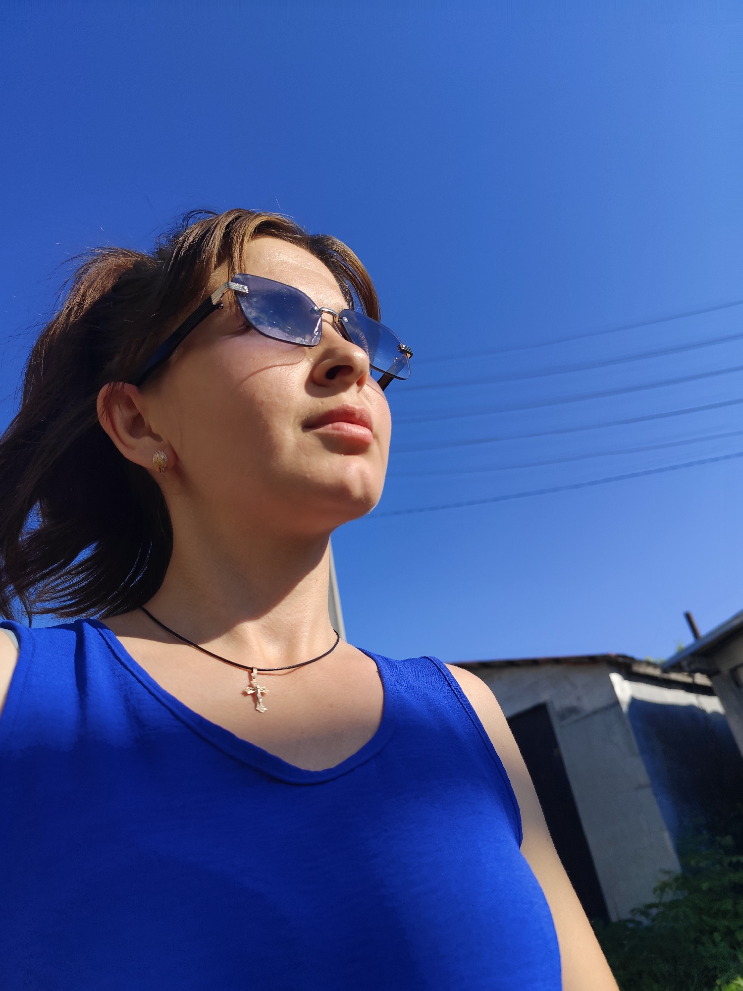 A woman wearing a blue tank top and sunglasses looks up at the sky. She has a cross necklace on. The background shows a clear blue sky, power lines, and buildings. The scene is located in the city of Fokino, Russia.