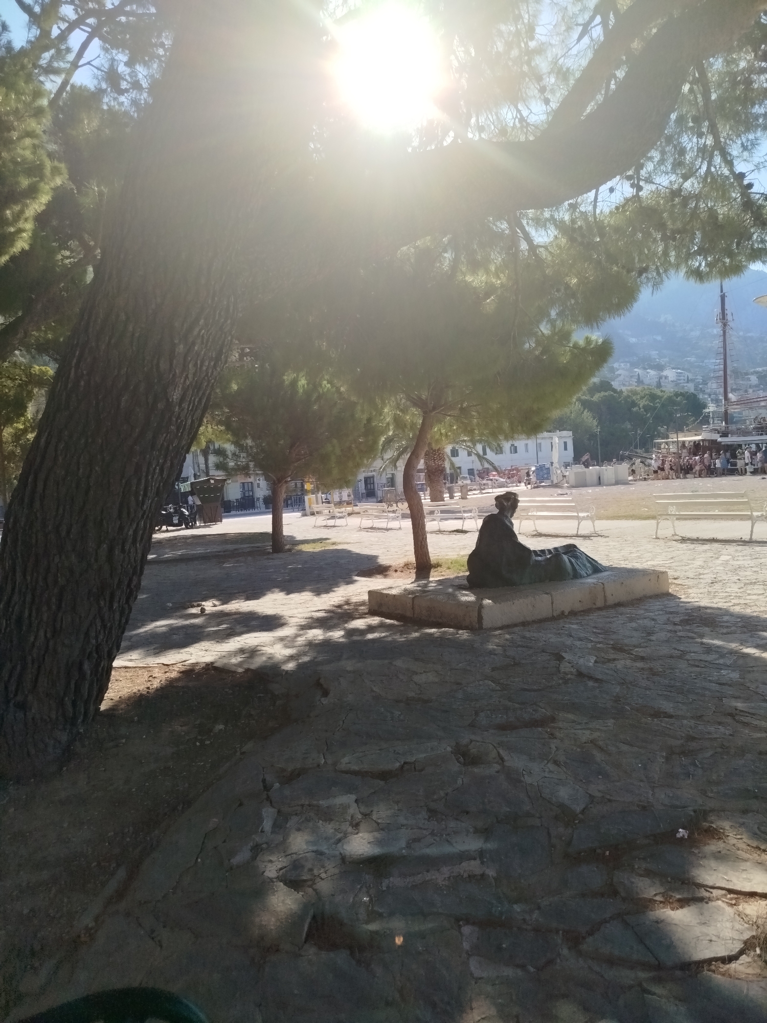 The image depicts an outdoor scene with a statue of a person sitting down. The statue is dark green and rests on a stone pedestal in a plaza. In the foreground, a large tree trunk is visible on the left, with its branches casting shadows on the stone-paved ground. Sunlight streams in from the upper right, creating a bright lens flare and illuminating the scene.
In the background, there is a body of water, likely a harbor, with boats docked. A promenade or walkway runs along the water, lined with white benches. A crowd of people can be seen on the promenade, some standing and others appearing to be walking. Buildings and trees are visible on the hillside in the far distance, suggesting a coastal town or city. The overall atmosphere appears to be calm and pleasant, possibly during a sunny day. The time of day is likely late morning or early afternoon, given the angle and intensity of the sun. There is no visible text in the image.