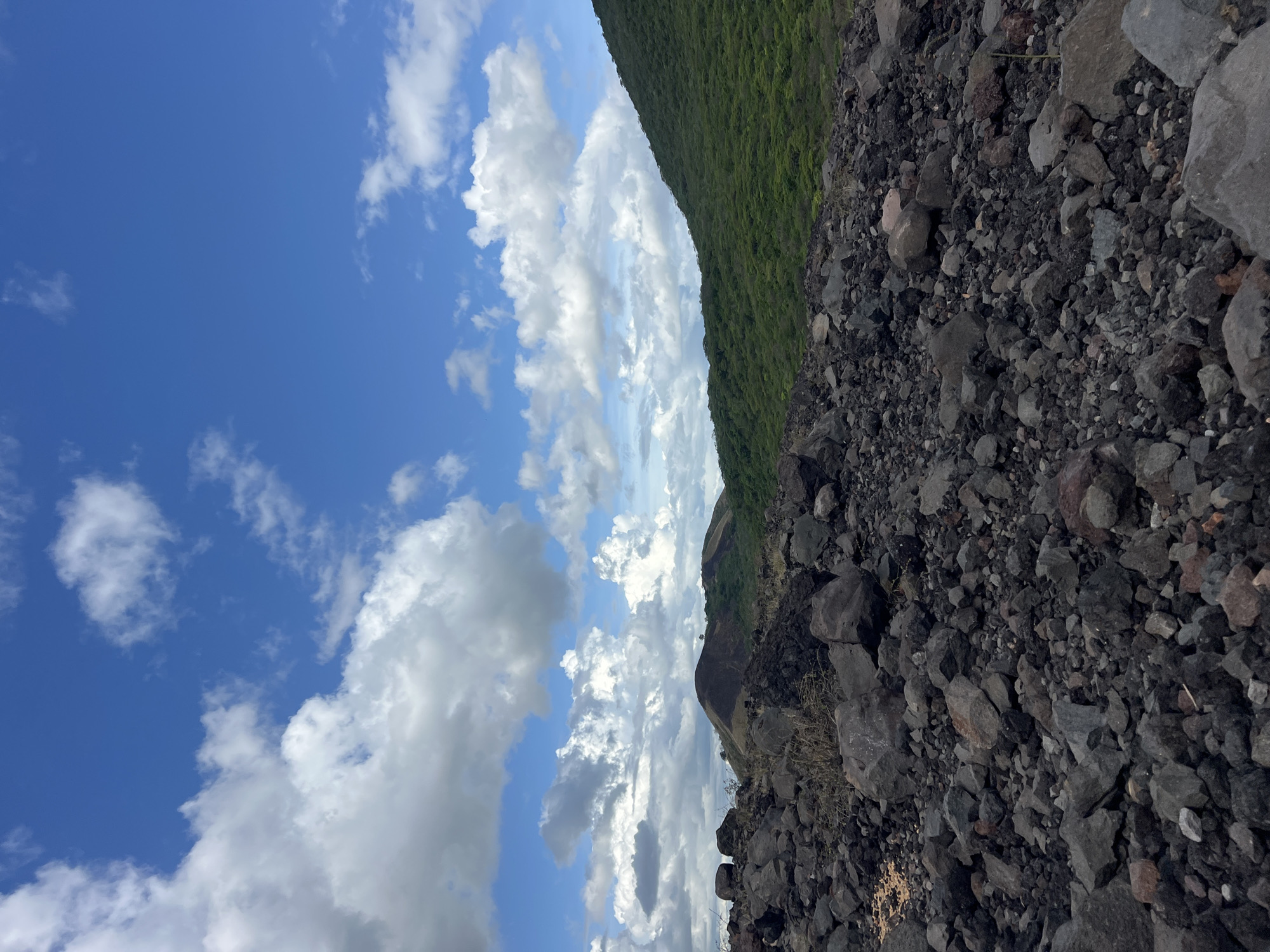 The image shows a rocky, volcanic landscape under a bright blue sky with fluffy white clouds. In the foreground, a dense field of dark, rough-hewn rocks fills the lower portion of the frame. Beyond the rocks, rolling green hills rise, covered in lush vegetation. In the distance, a dark, conical peak, possibly a volcano, is visible against the horizon. The scene appears to be an outdoor natural environment, suggesting a location like the volcanic plains or slopes near Rota, Nicaragua. There are no people or animals present in the image, and no specific actions or interactions are depicted. The overall impression is one of rugged, natural beauty and geological activity.