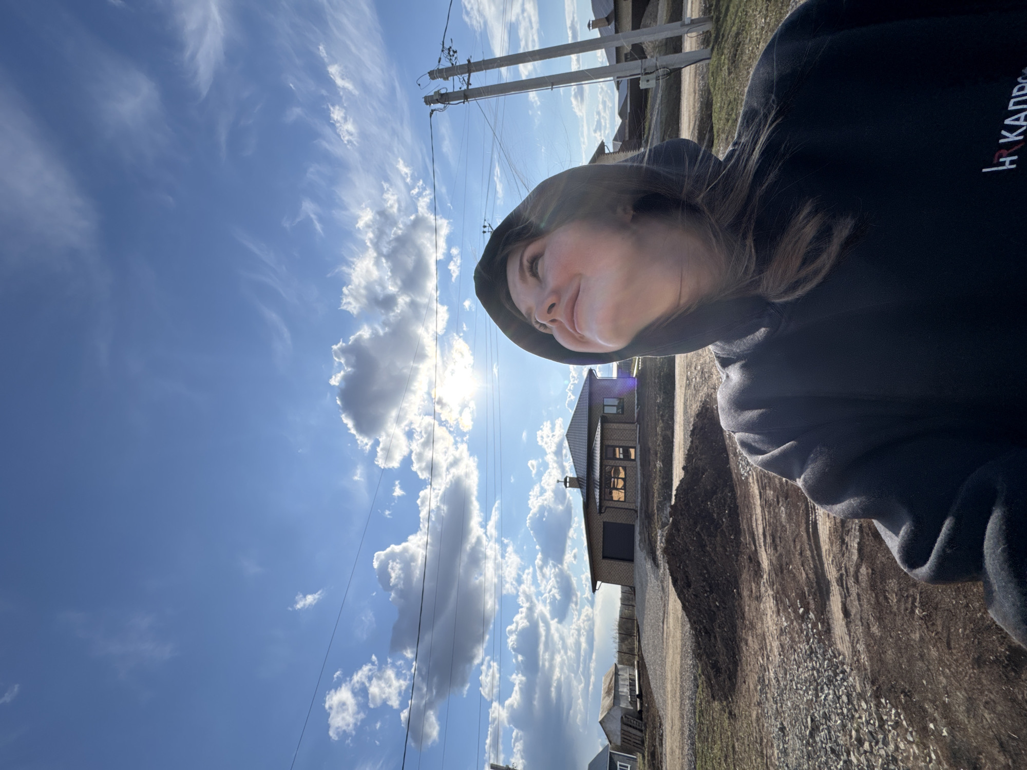 The image captures a bright, sunny outdoor scene in Gornyi (Dvurechenskoe S/P), Russia, featuring a young woman in a selfie or portrait-style shot. The woman, with long brown hair, is wearing a black hooded sweatshirt and is positioned in the right foreground, looking upwards towards the sun with a tranquil or slightly smiling expression. Her face is partially lit by the bright sunlight.
The dominant feature of the scene is the clear blue sky, which fills most of the frame, adorned with scattered white clouds ranging from wispy cirrus to more defined cumulus. The sun is visible as an intense bright spot in the upper left-middle, creating a lens flare. Utility poles with multiple wires stretch across the mid-ground, suggesting an area with infrastructure.
In the background, on the right, several buildings are visible, including a prominent house with a dark roof and a light-colored, possibly brick, facade. The ground in the mid-right and foreground is unpaved, appearing to be dirt and gravel.
A notable detail is the text "HR КАДРЫ" on the front of the woman's black hoodie, with "HR" in red and "КАДРЫ" (Russian for "Personnel") in white. The overall impression is a peaceful moment captured on a clear day in a rural or semi-rural setting.