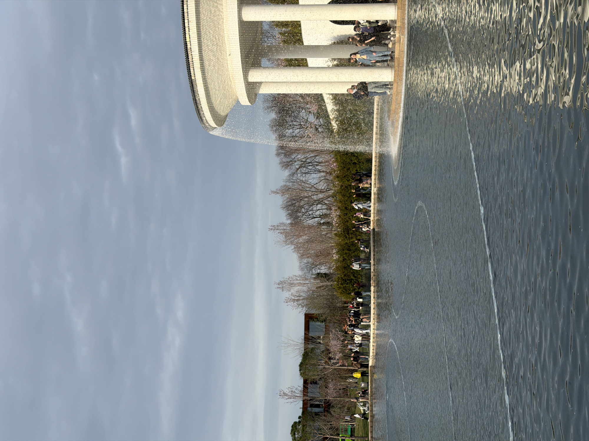 The image shows a public fountain with a curved architectural structure in Krasnodar, Russia. The fountain features a ring of water cascading down into a pool. People are seen sitting and standing around the edge of the fountain. The surrounding environment includes trees, grass, and buildings. The sky is overcast.