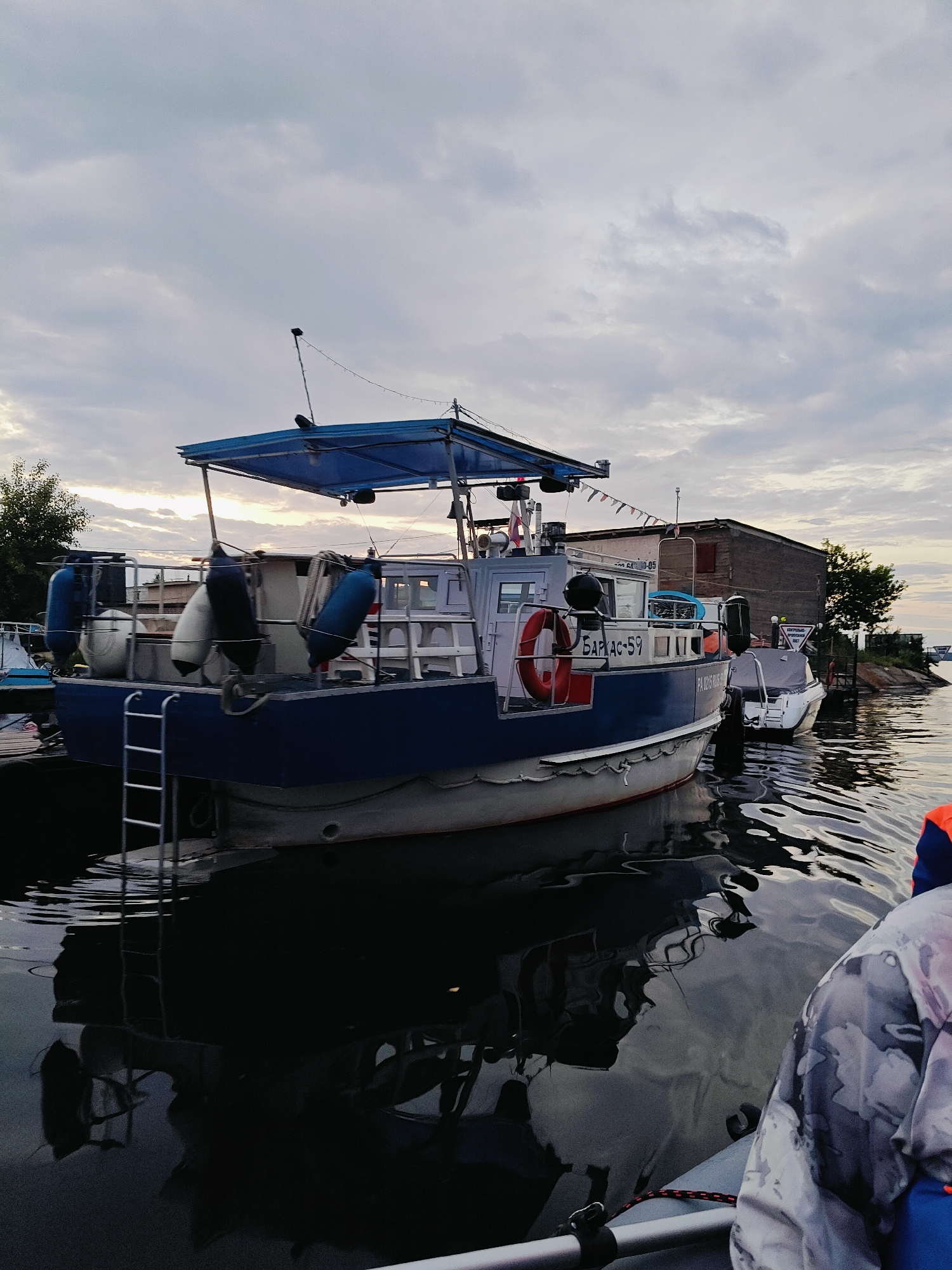 The image shows a cloudy sky over a body of water, likely a river or reservoir, in Chaikovsky, Russia. A blue and white boat is docked, with a ladder extending from its side to the water. Another smaller white boat is tied to the larger vessel. In the background, a weathered building and trees are visible along the shore. The water reflects the boats and the sky, creating a serene, subdued atmosphere. The image is captured from the perspective of someone in a boat looking towards the docked vessels, with a person wearing a grey and white jacket visible in the foreground.