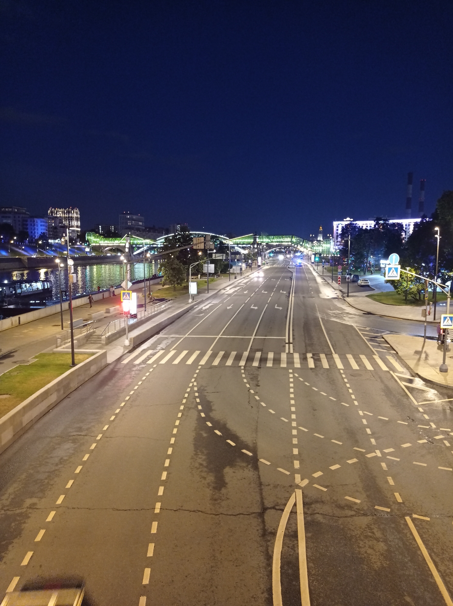 The image shows a wide, empty street in Moscow at night. The street is wet, likely from rain, with reflections of streetlights and building lights shimmering on its surface. Several pedestrian crossings with white stripes are visible. Traffic lights and directional arrows are painted on the road. On the left side of the image, a riverbank is visible, with a promenade and some trees. Buildings with illuminated windows and decorative lights are in the background, along with bridges crossing the river. The sky is dark blue, indicating it is nighttime. The scene appears to be on a major city artery, possibly a waterfront highway, given the presence of the river and the extensive infrastructure. There are no visible vehicles or significant human activity in the immediate foreground or on the street itself. A few people are walking on the riverbank promenade. The overall impression is one of urban stillness and quiet, characteristic of a city at rest after dark.