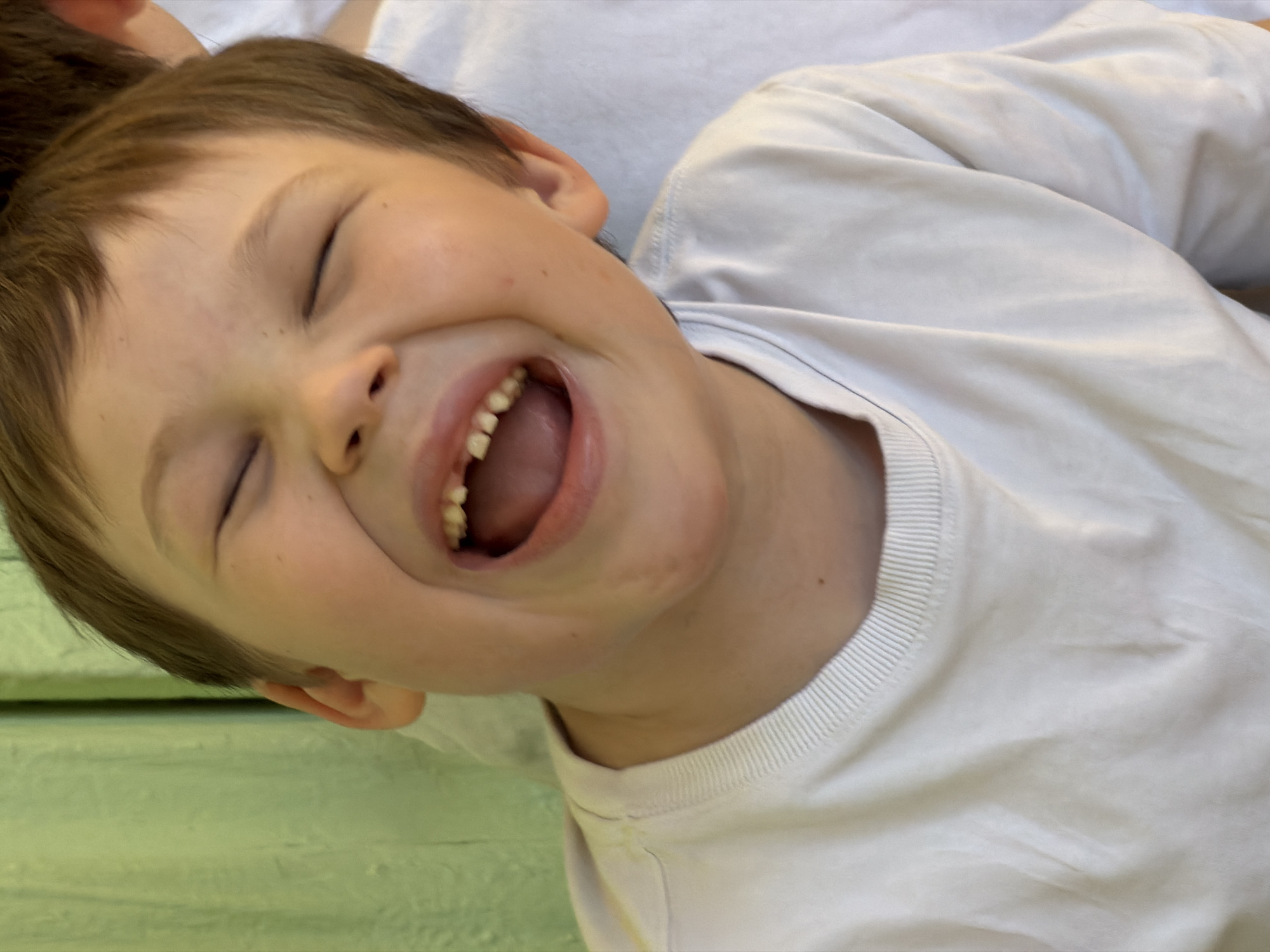 A close-up, tilted shot shows a young boy laughing with his eyes closed. He has light brown hair and is wearing a white shirt. His mouth is open, revealing his teeth and tongue. Part of another person's head is visible in the top left corner of the frame. The background includes a light green surface with some texture. The location of this scene in Izhevsk, Russia is not discernible from the provided image.