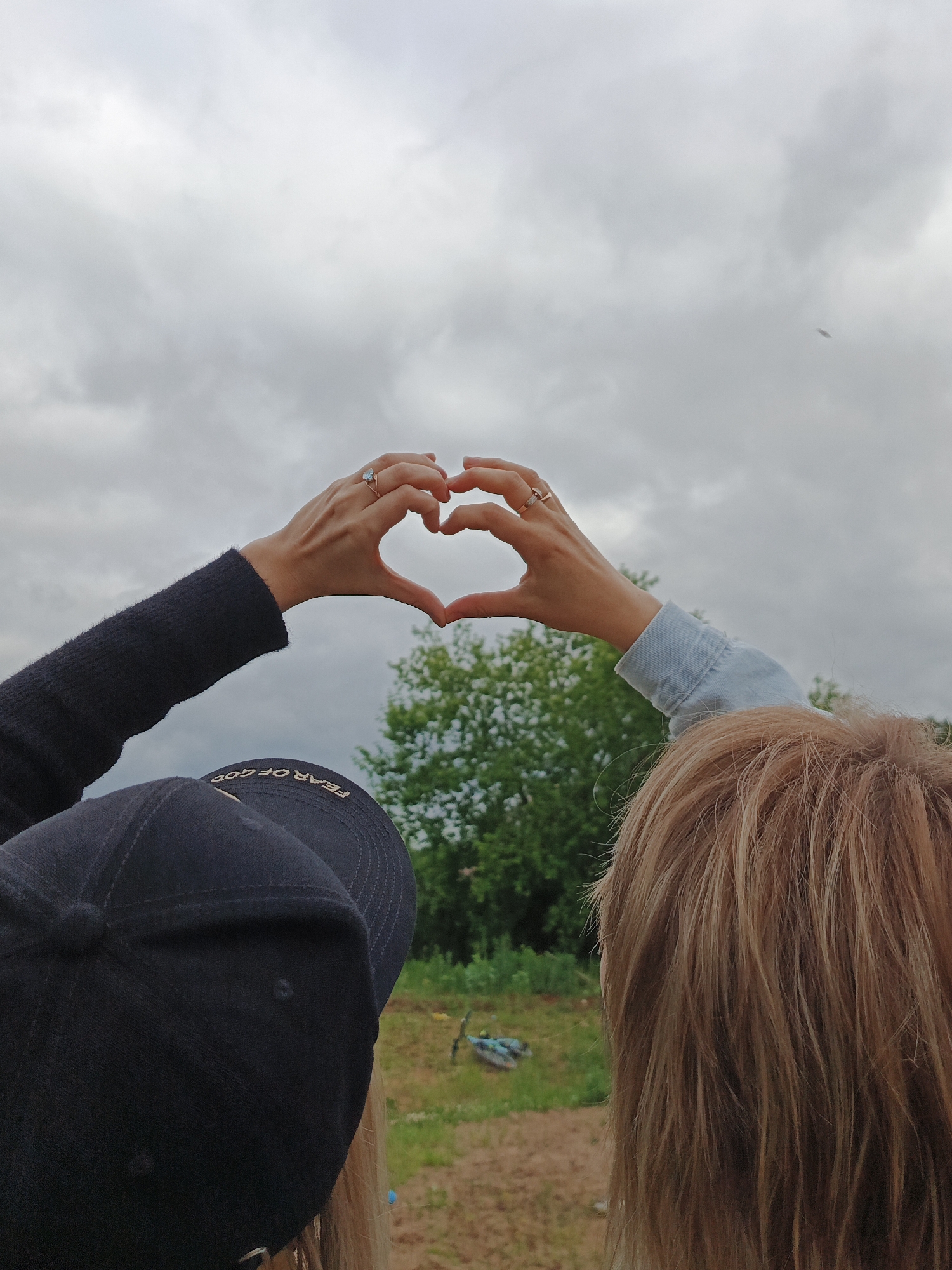 Two individuals are viewed from behind, outdoors under an overcast sky. They are forming a heart shape with their joined hands in the upper center of the frame. The person on the left wears a black baseball cap with "FEAR OF GOD" in gold lettering visible upside down on the brim, and a long-sleeved black garment. Their left hand, adorned with a gold ring featuring a light blue stone on the ring finger, contributes to the heart gesture. The person on the right has shoulder-length, light brown hair and wears a light blue denim sleeve. Their right hand, displaying a gold-toned ring with small embedded details on the ring finger, completes the heart. The background features a dense line of green trees and foliage directly behind the individuals' hands. The ground below is a mix of light brown earth and scattered green grass. A blue and black bicycle lies horizontally on the ground in the mid-distance to the right. The sky above is uniformly gray and heavily clouded. A small, dark, indistinct object is visible in the upper right portion of the sky.