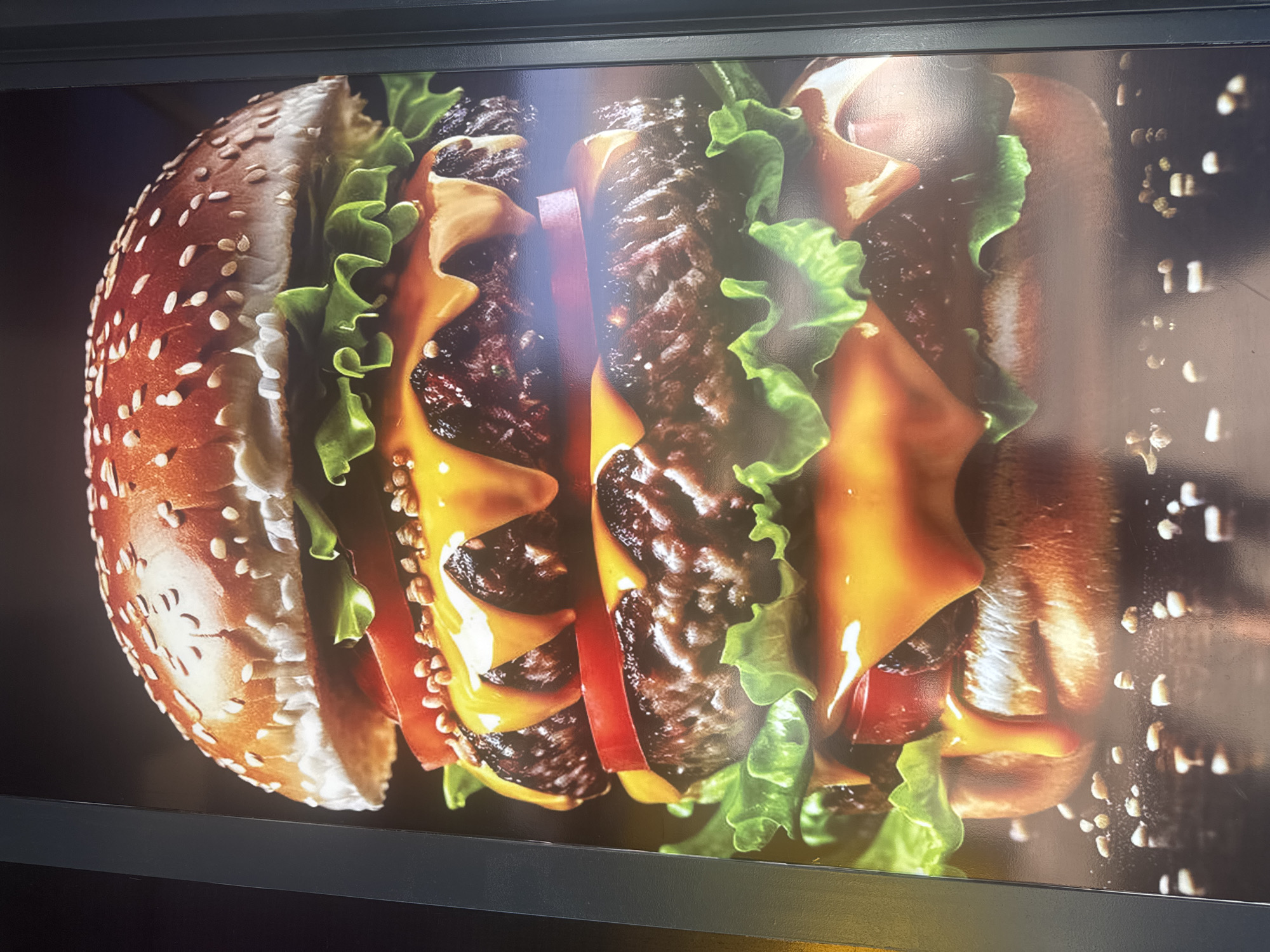 The image depicts a close-up of a double cheeseburger with sesame seeds on the bun, lettuce, tomato, and melted cheese. The photo is likely a poster or advertisement displayed in a restaurant or fast food establishment. The location of the photograph is unknown as there is no location information in the image.