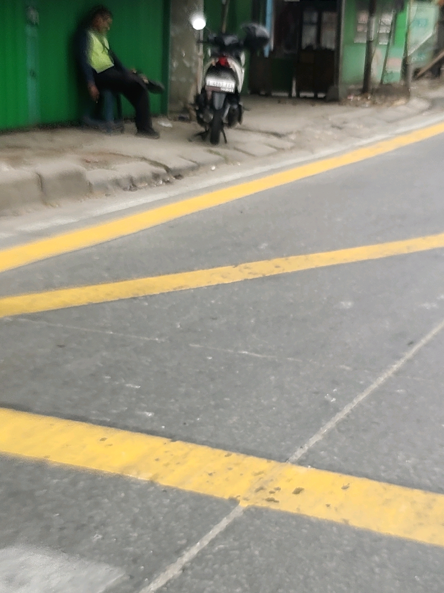 A person is leaning against a green wall in the city of Mekarmukti, Indonesia. The person is wearing a yellow vest and black pants. They appear to be resting or sleeping. In the background, there is a motorcycle parked next to the wall. The ground in front of the wall is a sidewalk with some debris. A road with yellow lines runs in front of the sidewalk. The overall scene depicts a street in a somewhat urban environment.