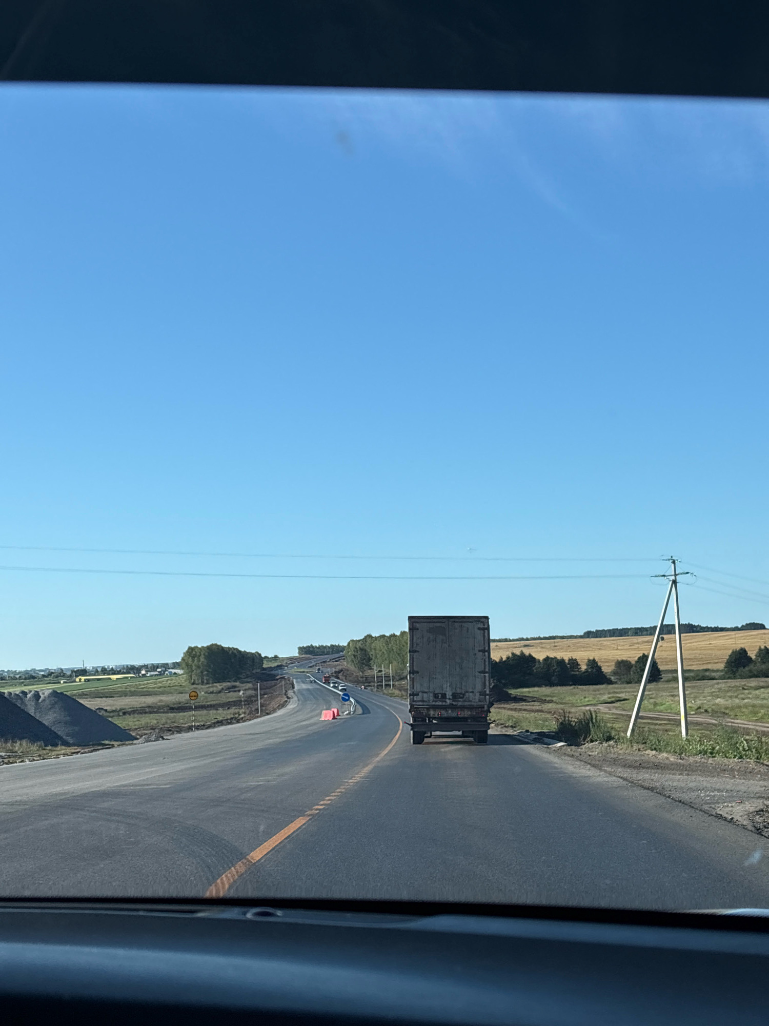 A forward-facing perspective from inside a vehicle shows a two-lane highway under a clear blue sky. The road curves to the right in the distance. On the left side of the road, piles of gravel and construction barriers are visible, indicating roadwork. A large, gray truck with a closed trailer is driving ahead in the right lane. In the background, fields of dry grass and trees border the highway, with a power line and pole on the right. A small town or cluster of buildings is visible on the far left. The scene appears to be in a rural area.