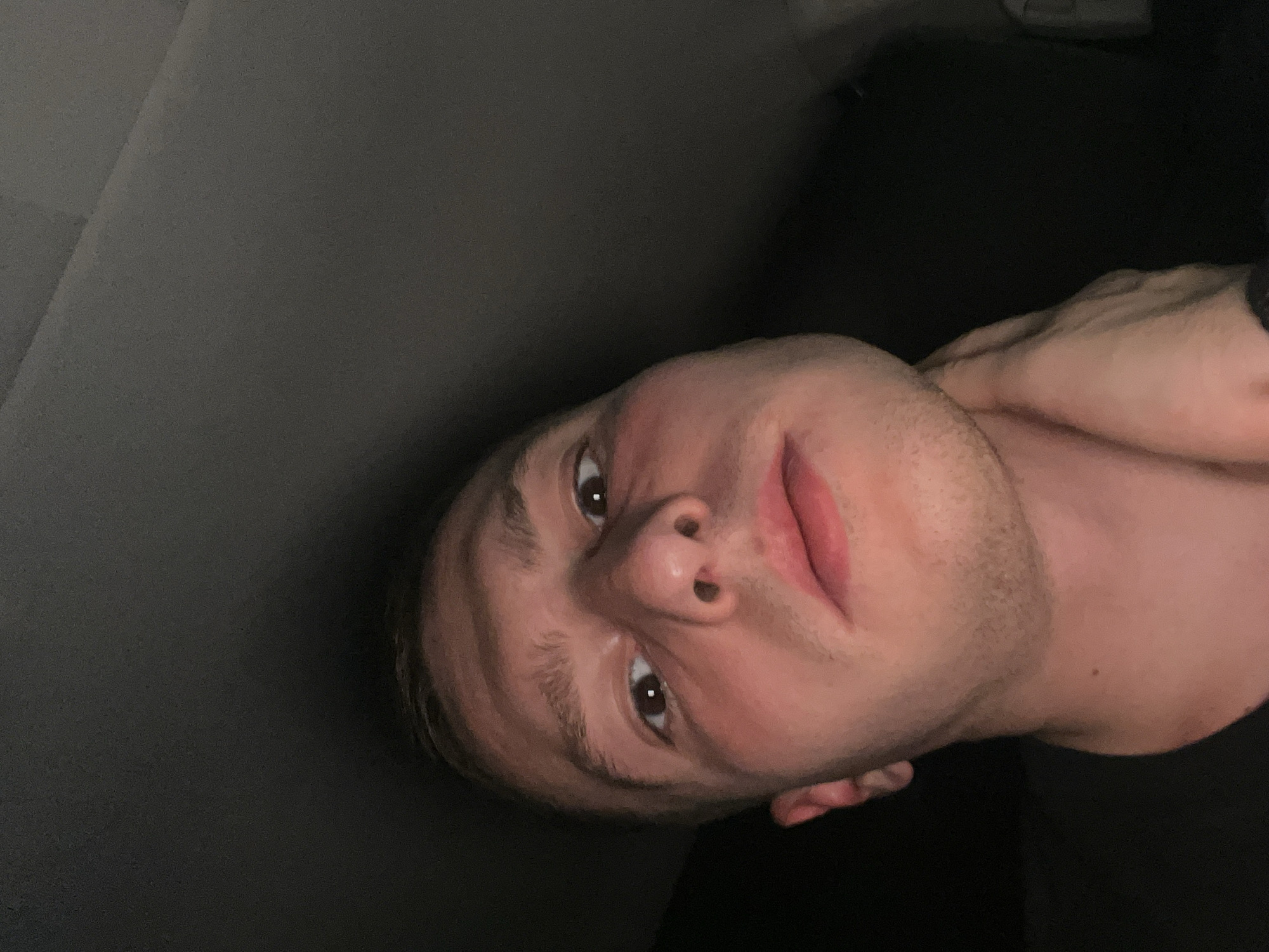 The image is a selfie of a young man with light skin. He has brown eyes, short light brown hair, and light stubble on his face. His expression is neutral. He is wearing a dark shirt. He is holding his hand to his neck. The background is dark, but a light gray surface can be seen. It is difficult to determine the location in Burgas, Bulgaria, as there are no identifiable landmarks or details visible in the image.