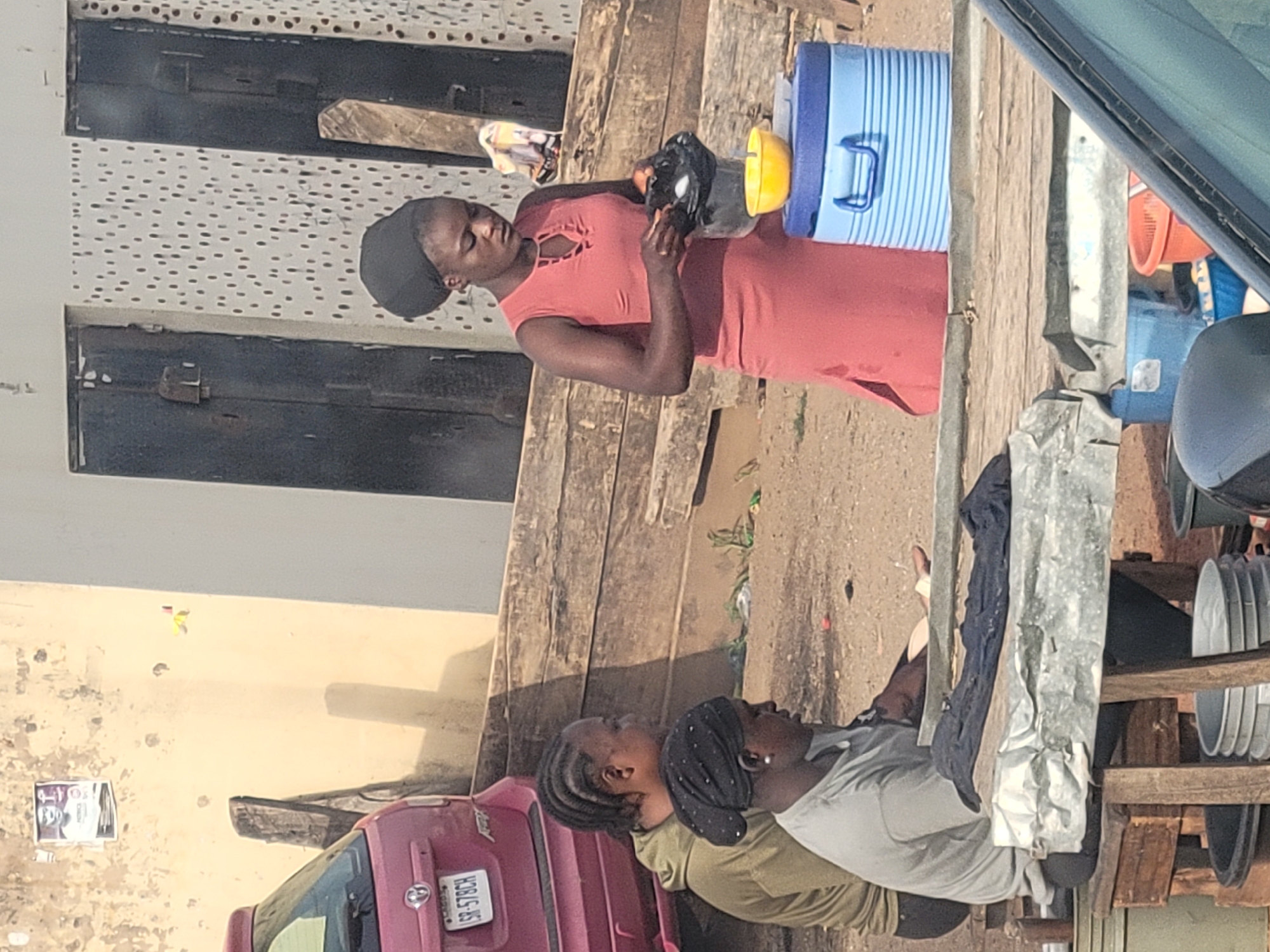 The image shows a street scene in Jos, Nigeria. A woman in a red dress and black head covering is filling a black and yellow container from a blue and white cooler. Nearby, another woman sits on the ground next to a pink car with license plate "SR-578CH". Clothes hang to dry on a wooden structure with metal support. Dishes are stacked on a rack. The background includes buildings with textures, including a white building with dark spots and a tan wall with a poster.