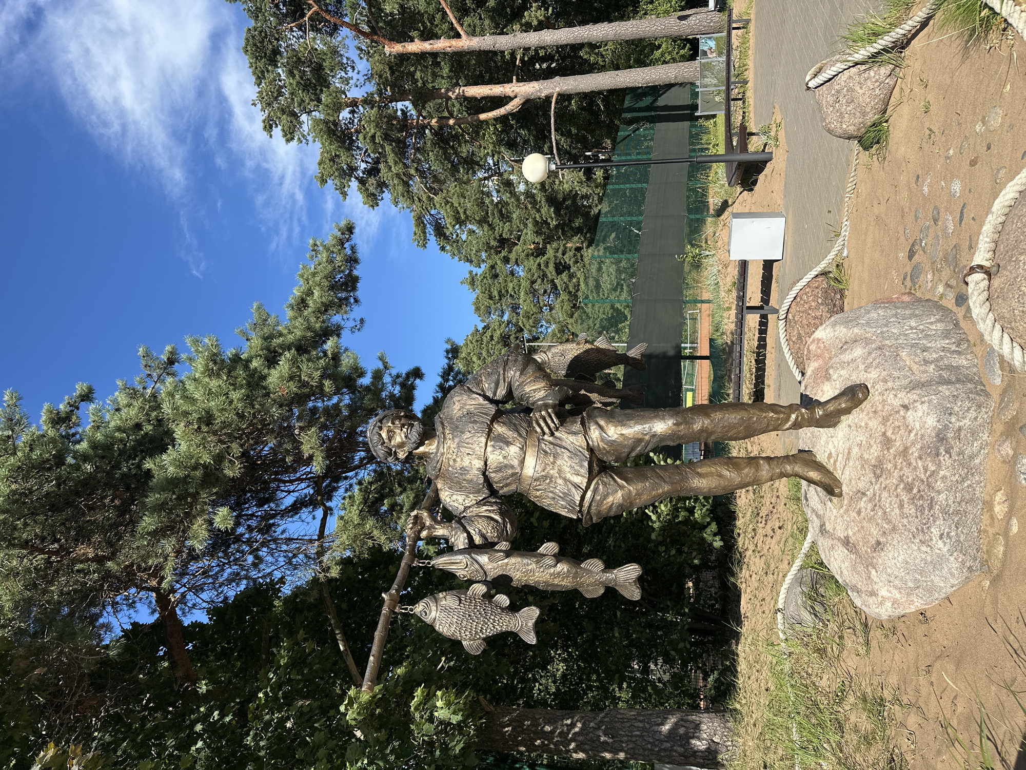 The photograph captures a bronze statue of a fisherman in the city of Zelenogorsk, Russia. The fisherman, depicted in historical attire, stands on a large boulder, holding a fishing rod over his shoulder. Three fish are suspended from the rod, indicating a successful catch. The statue is situated outdoors, surrounded by lush green trees and a bright blue sky with scattered white clouds. In the background, there are tennis courts with a green fence and seating areas, suggesting a recreational park or sports complex. The sandy ground around the statue is decorated with large rocks and thick ropes, adding a nautical theme to the scene. The lighting suggests a sunny day, casting shadows on the ground and highlighting the metallic sheen of the statue.