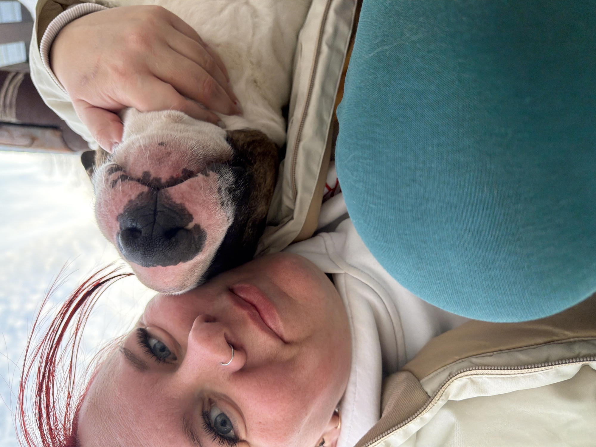 The image displays a close-up, low-angle view of an adult female embracing a dog, set against a cloudy sky in Chaikovsky, Russia. The female, positioned on the left and center, has light skin, blue eyes with prominent dark eyelashes, and a silver hoop nose ring in her left nostril. Streaks of reddish-orange hair are visible on the right side of her head. She is wearing a light-colored hooded sweatshirt underneath a beige zippered jacket. A teal garment, likely a pant leg, is visible on the far left. Her right hand is seen holding the dog's jowl area. The dog, a brachycephalic breed consistent with a Bulldog, has white and brown/brindle fur on its face, a dark black nose, and visible pink skin around its snout and mouth. Both the female and the dog are oriented towards the camera. The background consists of a partially cloudy sky, with blurred dark brown and striped architectural elements faintly visible on the far right.