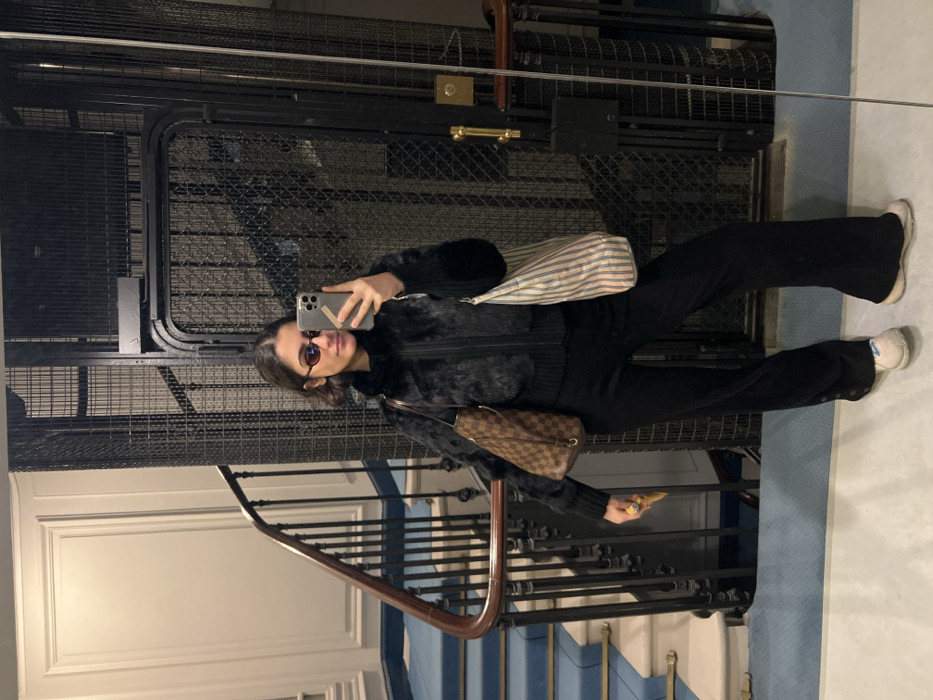 The image displays a young woman taking a mirror selfie within the entrance hall or stairwell of a building in Paris, France. She has short, dark hair and wears dark, oval-shaped sunglasses. Her attire includes a black faux fur zippered jacket, black wide-leg trousers, and light-colored athletic sneakers. In her right hand, she holds a smartphone, actively used to capture the image, while her left hand holds a small yellow folded item. She carries two bags: a brown Louis Vuitton "Damier Ebene" pattern bucket bag on her left shoulder and a vertically striped fabric tote bag on her right shoulder. The environment exhibits classic Parisian architecture, featuring a white wall with ornate molding, a dark wooden staircase with metal balusters and blue carpeted steps on the left, and a vintage black metal grille elevator with a brass handle on the right.