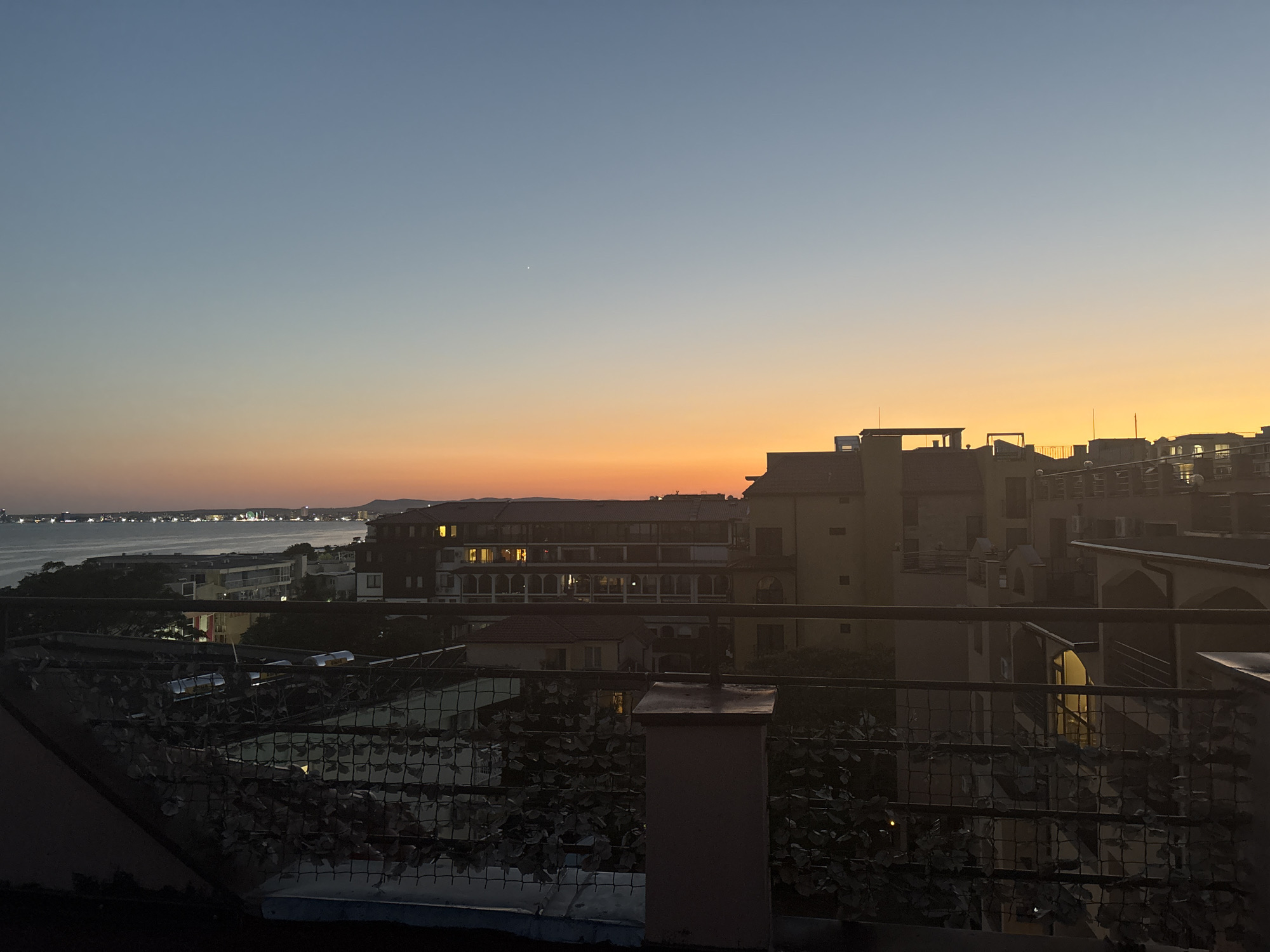 The image captures a twilight view of a coastal city, likely Sveti Vlas, Bulgaria, from an elevated vantage point. The sky transitions from a clear, pale blue at the top to a vibrant gradient of orange and yellow near the horizon, indicating the sun has recently set. A faint, white star-like object is visible in the upper portion of the sky.
In the middle ground, a body of water, likely the Black Sea, stretches horizontally across the frame. Along the coastline, buildings of varying architectural styles are clustered together. Some buildings are illuminated from within, with windows emitting a warm, yellow light, suggesting it is evening. The architecture appears to be a mix of modern and traditional European styles, with some buildings featuring balconies and multiple stories.
In the foreground, a dark metal railing with a decorative pattern runs across the bottom of the image, partially obscuring the view of the city and water. Some artificial foliage is attached to the railing, adding a touch of texture and color. The overall atmosphere is serene and peaceful, characteristic of dusk in a coastal town. No people are visible in the image.