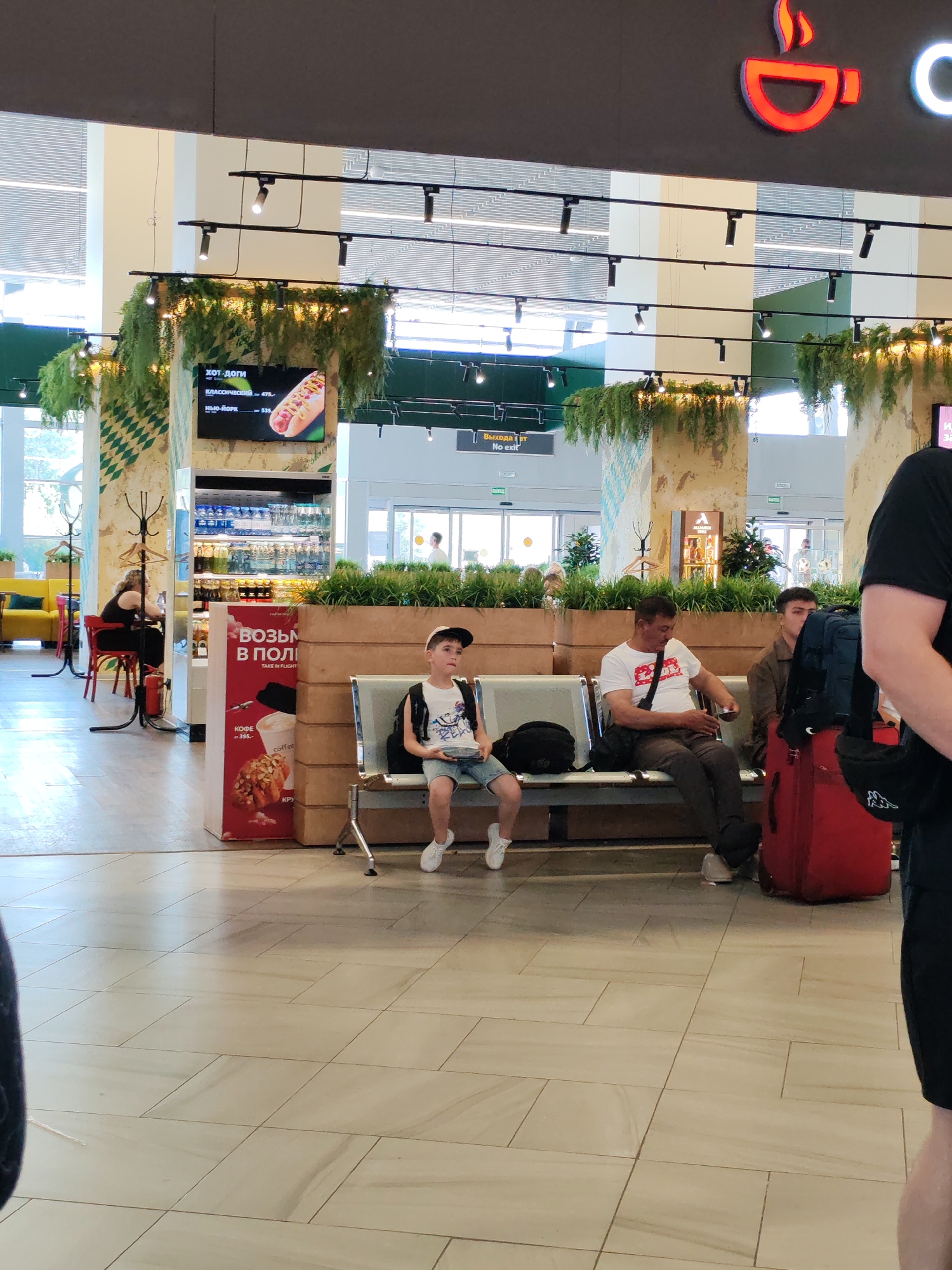 The image captures a scene inside an airport terminal, likely within Khrabrovo Airport, Russia, given the context provided. In the foreground, a young boy wearing a white t-shirt, blue shorts, a baseball cap, and white sneakers sits on a metallic bench, holding a small white plate. He is facing slightly to his right, looking towards the right side of the frame. To his right, an adult male in a white t-shirt with red lettering and dark pants is seated on the same bench, looking down at something in his hand. To the right of this man, another young male wearing a brown jacket sits on the bench, looking towards the front. Between the seated individuals and the right edge of the frame, a large red suitcase is visible. Behind the bench, a low planter with green foliage is situated. In the background, a refrigerated display case is stocked with various drinks, and above it, a digital menu board displays images of hot dogs and their prices. A coffee cup logo is visible on the upper right of the main image. Further in the background, the terminal features large glass doors, some seating areas, and promotional displays. The overall environment suggests a public waiting area in an airport.