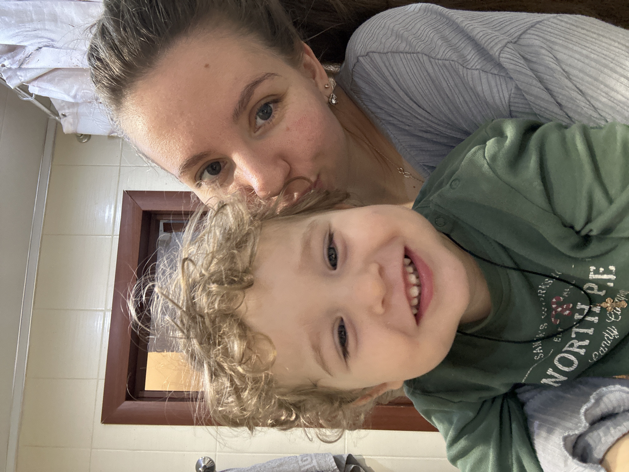 In an indoor setting in Klin, Russia, an adult female is depicted holding and kissing a young male child. The female, positioned behind the child, has light skin, visible dark hair pulled back, and a light blue left eye. She wears a light grey ribbed long-sleeved shirt, a delicate gold-toned necklace with an infinity symbol, and a small stud earring in her left earlobe. Her lips are pressed against the child's right cheek. The child, appearing to be a toddler, has light skin, curly blonde hair, and is smiling broadly towards the camera, revealing his upper and lower front teeth. He wears a dark green long-sleeved shirt with the partial inscription "SANTA'S WORKSHOP NORTH POLE" and a black cord necklace featuring a small gold-toned cross pendant. The background consists of light cream square tiles with dark grout lines. A dark brown wooden door frame is visible on the left, next to a silver-colored fixture from which a grey towel with a geometric pattern hangs. On the right, a white pleated shower curtain is visible, suspended from a silver-toned rod. The scene is brightly and evenly lit.
