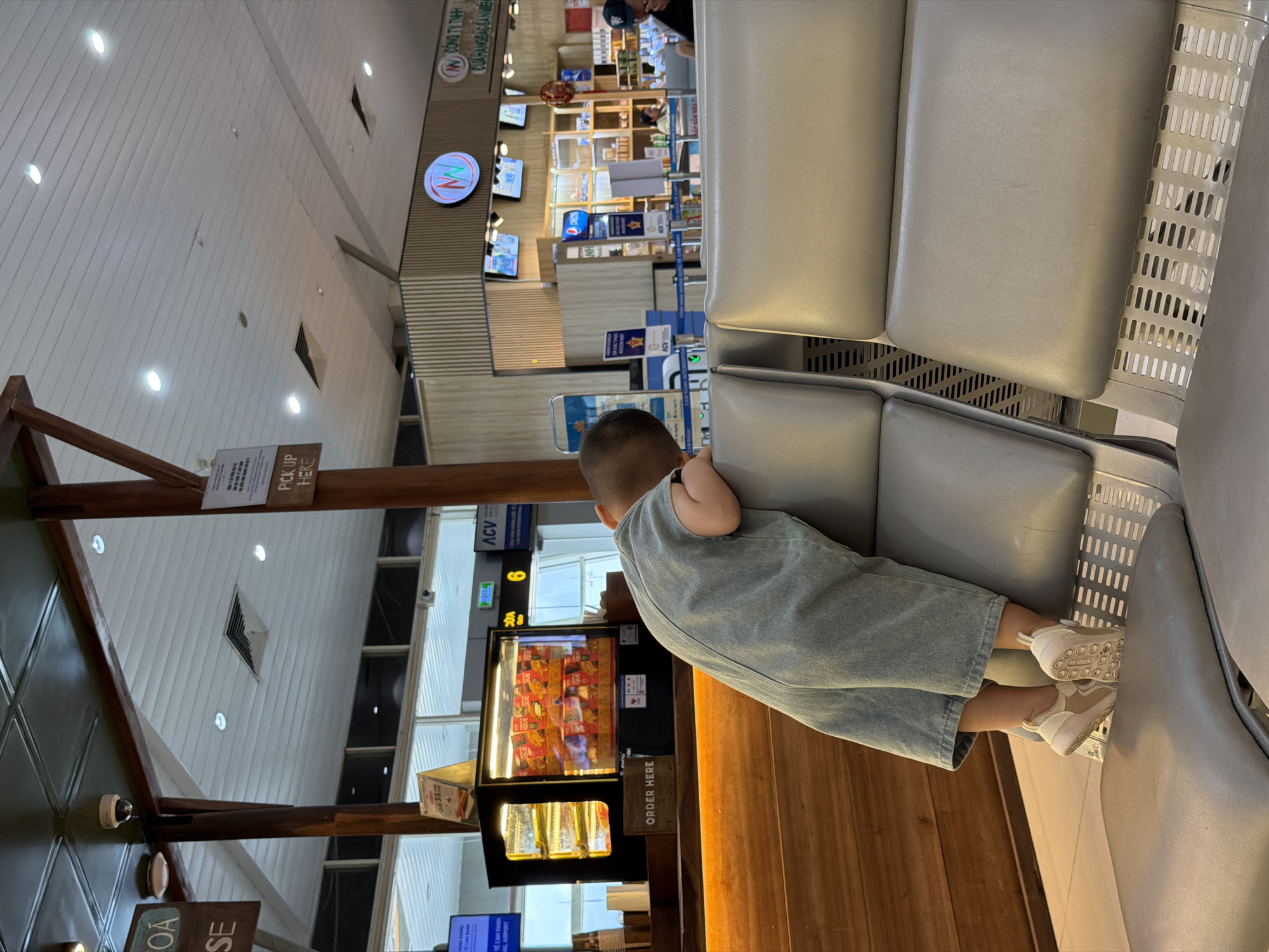 In a waiting area, possibly at Cam Ranh International Airport, Bac Cam Ranh Ward, Vietnam, a young child in a light blue denim romper stands on a grey airport seat. The child is leaning forward, with their head resting on the back of the adjacent seat. Their light-colored shoes are visible, pressing against the seat. The background shows a retail area with a refrigerated display case filled with snacks and a counter with an "ORDER HERE" sign. Beyond that, a shop displays various products on shelves. The overall environment appears to be a public transportation hub, characterized by seating arrangements and commercial establishments.