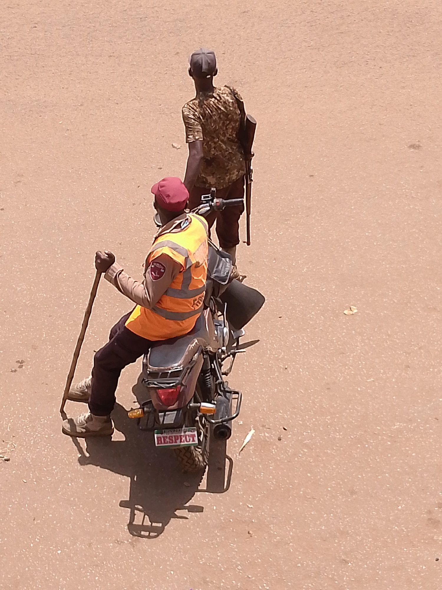 The image shows two men on a paved road in Bukuru, Nigeria. One man sits on a motorcycle, wearing a red cap, an orange vest with patches, and dark trousers. He is holding a wooden stick. The motorcycle's license plate reads "RESPECT." The other man is standing behind the motorcycle, wearing a camouflage shirt, dark trousers, and a dark cap. He has a rifle slung over his shoulder. The road is tan in color. The image is taken from an elevated perspective.