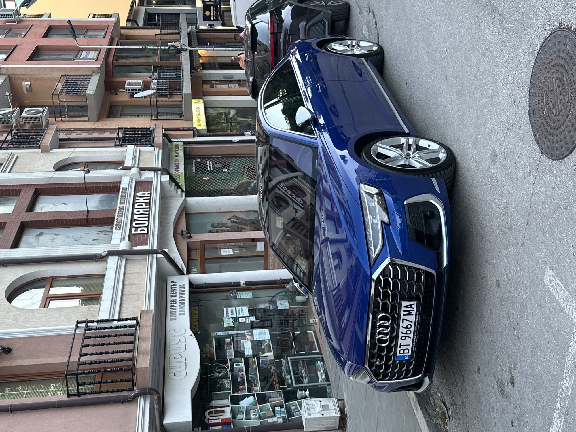 This image shows a street scene in Veliko Tarnovo, Bulgaria. The foreground is dominated by a blue Audi S8 parked on the street. The car has a license plate that reads "BT 9667 MA". Behind the Audi, another dark-colored SUV is visible. The background consists of a row of multi-story buildings with shops on the ground floor. One of the shop signs, written in Cyrillic script, says "БОЛЯРКА" (Bolyarka), which is a popular Bulgarian beer brand, and likely a business name. Another sign reads "СИРУС" (Sirius) above a shop selling souvenirs. There are people visible on the balconies and in the shops. The street is paved, and a manhole cover is visible in the lower right corner. The overall scene depicts a typical urban environment in Bulgaria.