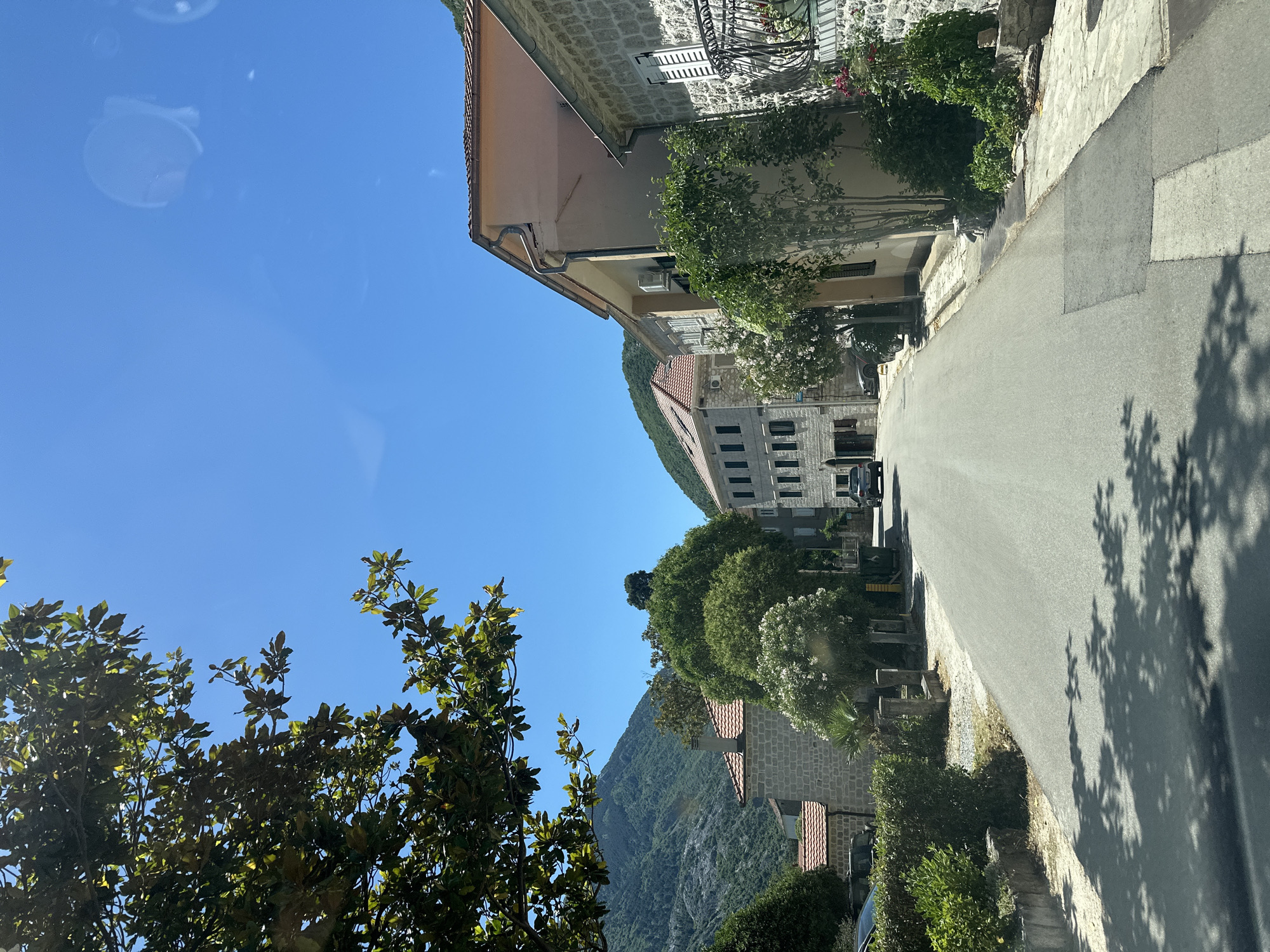 The image depicts a street scene in Muo, Montenegro, observed from a slightly elevated perspective. A paved road dominates the foreground, angling upwards towards the center of the frame. The road is marked by tree shadows and bordered on both sides by buildings and vegetation. The architecture consists of stone buildings with tiled roofs, and facades adorned with plants. In the background, a mountain rises into a clear blue sky. In the distance, vehicles can be seen parked along the street. The photograph is taken during daylight under clear skies.