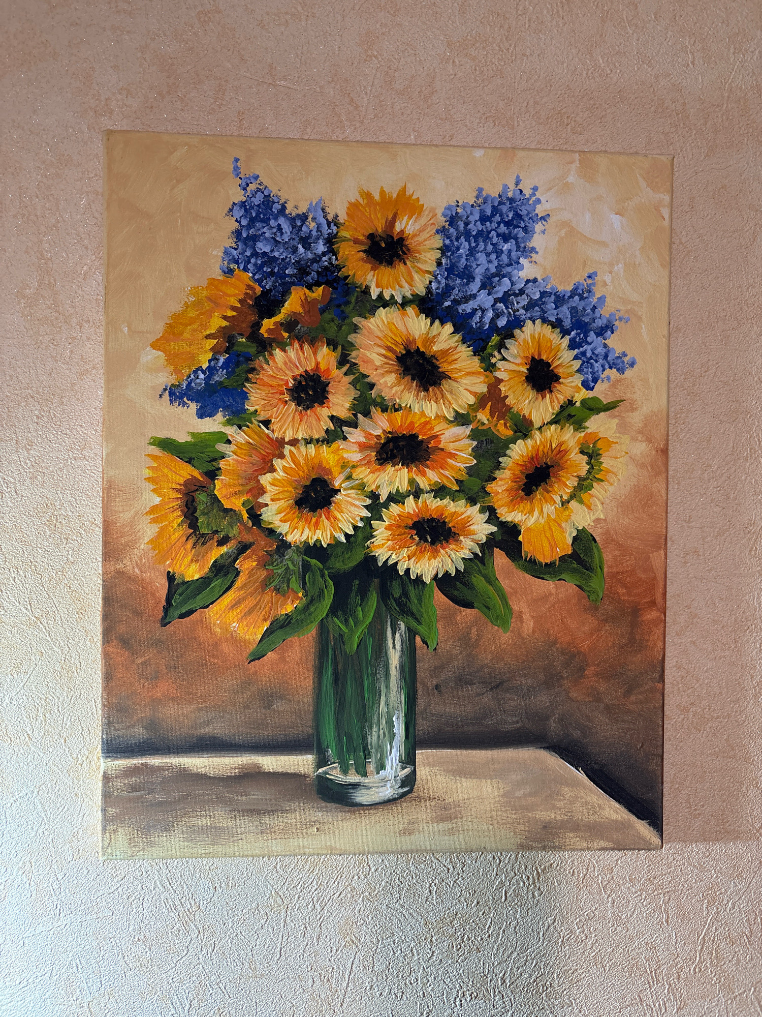 This is a forensic, matter-of-fact description of an image depicting a painting of a bouquet of sunflowers and blue flowers in a glass vase. The painting is displayed against a textured wall with a peach hue. The sunflowers are depicted with yellow petals and dark centers, arranged in a full bouquet. Blue floral elements are interspersed among the sunflowers. The vase is clear glass, showing green stems within. The painting rests on a surface that appears to be a table or shelf, rendered in shades of beige and brown with a shadow cast behind it. The overall impression is of a still life composition. There is no identifiable location in the city of Ternopil, Ukraine, or any people present in the image.