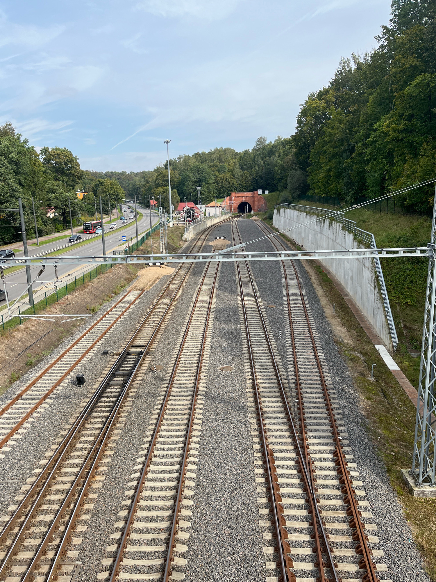 The image depicts a wide view of a railway junction and an adjacent highway, set against a backdrop of dense trees and a cloudy sky. The foreground is dominated by multiple sets of parallel railway tracks, with gravel ballast and concrete sleepers visible. In the middle ground, the railway lines converge towards a red brick tunnel entrance. To the left of the railway lines, a multi-lane highway is visible, with several cars and a red bus in motion. The highway is bordered by green fencing and mature trees. In the background, the trees form a dense green wall along a slope. The overall scene is one of urban infrastructure integrated with nature.
The location is identified as the railway tunnel in Kaunas, Lithuania. This specific tunnel is part of the Kaunas railway tunnel complex, which is a notable landmark and a significant piece of infrastructure in the city. The highway visible is likely part of the infrastructure surrounding the tunnel.