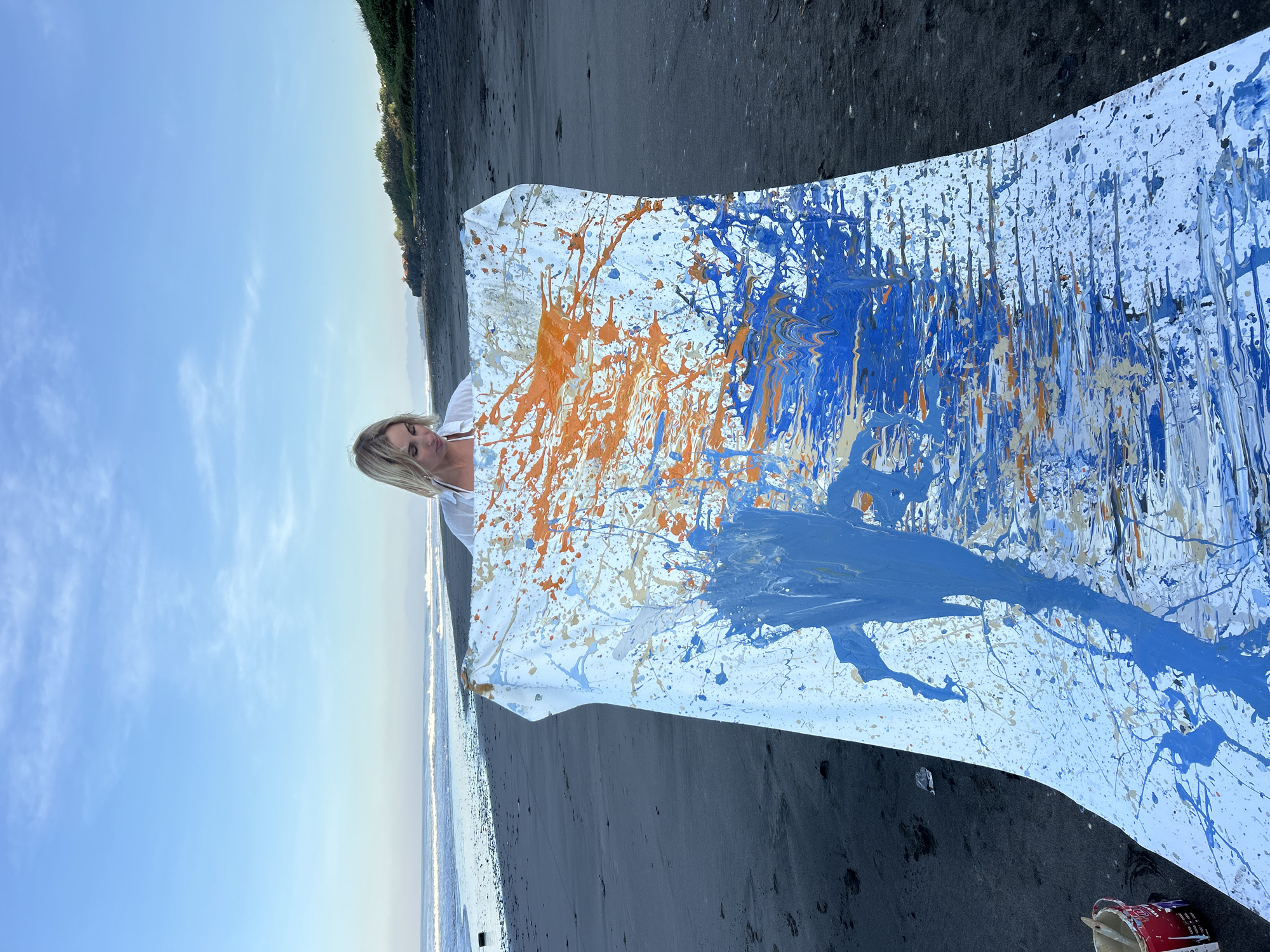 A blonde woman stands on a black sand beach in Tabanan, Indonesia, holding a large, white canvas covered in abstract blue and orange paint splatters. The canvas is unfurled, appearing to be a fluid piece of art. The woman wears a white, open shirt over a white bikini top. The ocean stretches out behind her with gentle waves breaking on the shore. In the distance, lush green vegetation lines the coast. The sky is a clear, pale blue with wispy clouds. To the left of the canvas, a red bucket containing paintbrushes is visible on the sand. The overall scene suggests an artist working outdoors in a scenic coastal environment.