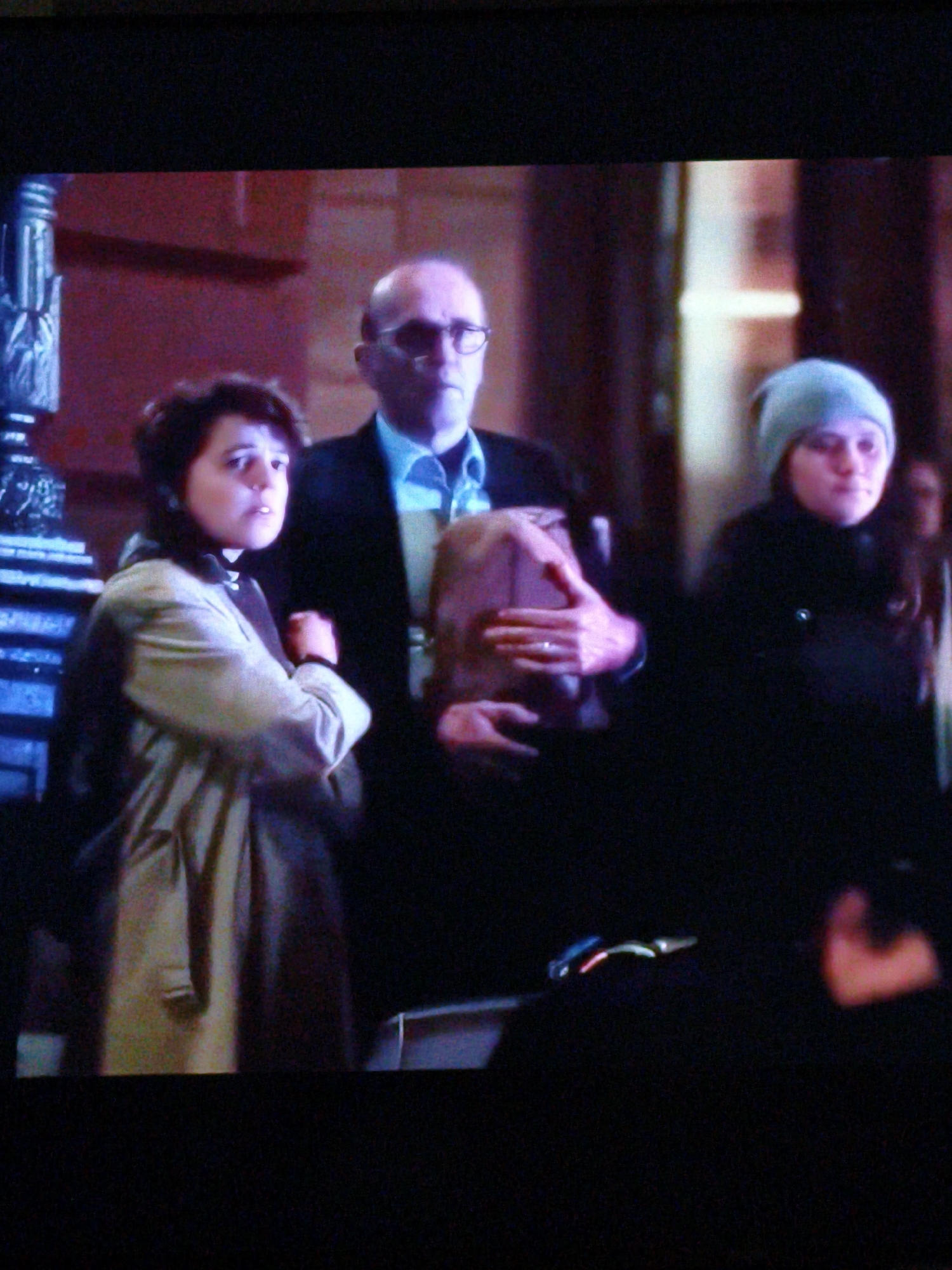 In a dimly lit indoor setting, three individuals are visible. On the left, a person with short dark hair wearing a light-colored trench coat with dark lapels has their arms crossed. In the center, a bald man with glasses, wearing a dark suit jacket and a light blue collared shirt, holds a brown object. To the right, a person wearing a light blue beanie and a dark jacket looks towards the camera. The background features muted red walls and a dark, indistinct structure on the far left. The overall scene appears to be captured from a distance, with low resolution and notable digital noise. There is no visual information in the image that indicates a connection to Vladivostok, Russia.