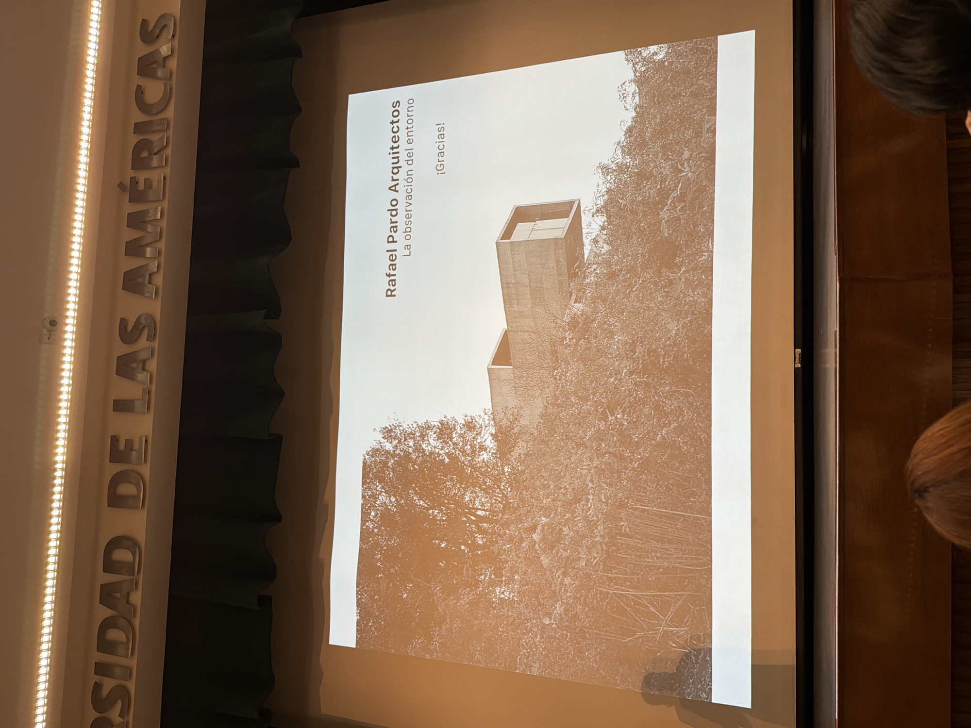 Here is a description of the image:
The image shows a projector screen displaying a presentation slide. The slide is titled "Rafael Pardo Arquitectos: La observación del entorno" ("The observation of the environment"), with the word "¡Gracias!" ("Thank you!") below. The background of the slide features a sepia-toned image of a modern concrete structure partially obscured by trees.
To the left of the screen, a vertical panel reads "RSIDAD DE LAS AMÉRICAS" in large, vertically stacked letters illuminated by a string of lights. The panel is set against a dark, curtain-like backdrop.
On the right edge of the image, parts of people's heads can be seen, suggesting that an audience is present. The overall scene appears to be a lecture or presentation setting, likely within a university context.