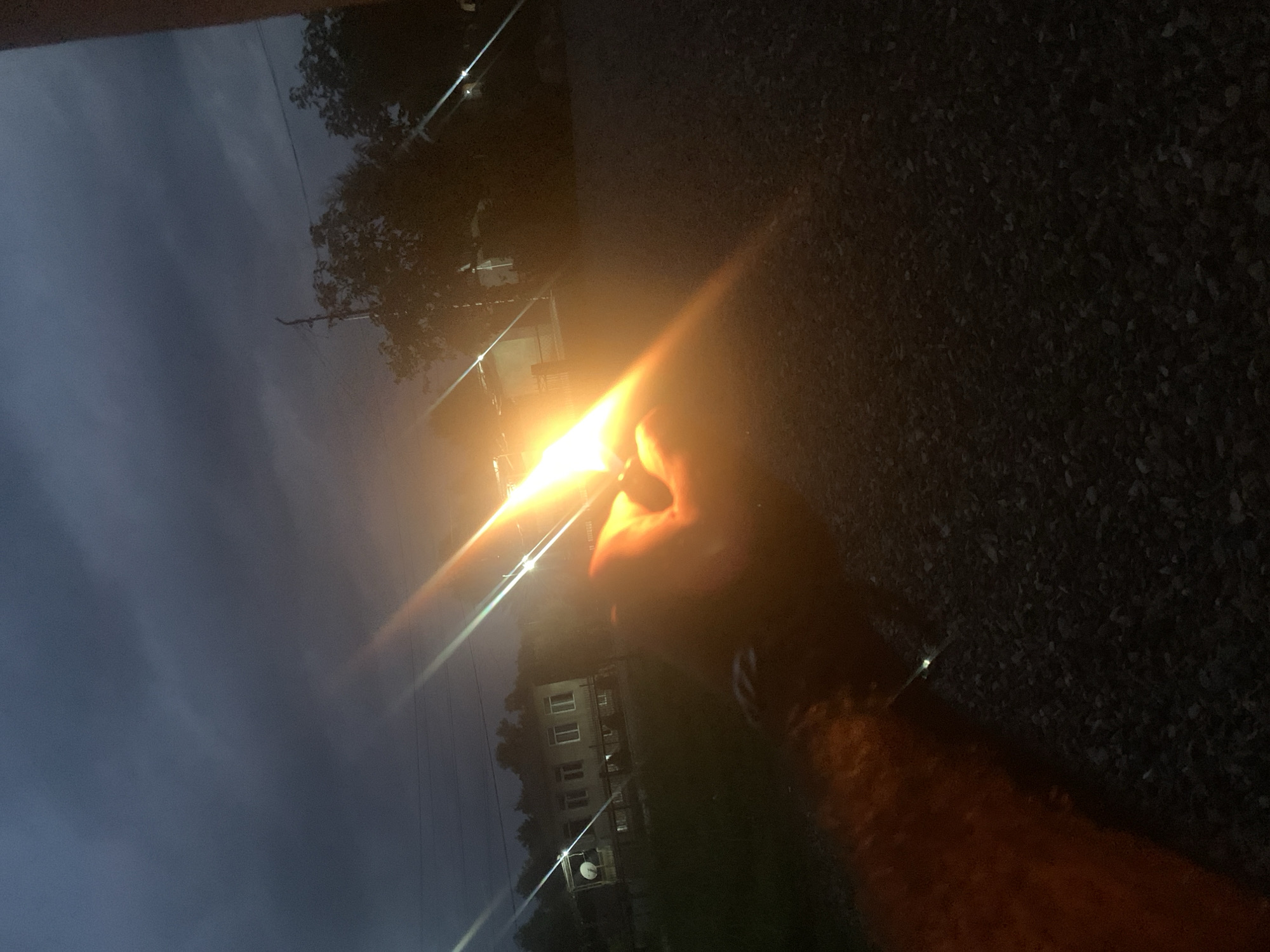 A hand is visible in the foreground, holding a lit lighter. The flame of the lighter is bright and casts a warm orange glow. The background is dark and out of focus, with hints of trees and buildings. The ground appears to be covered in gravel. The image has a dark, moody atmosphere, possibly taken at dusk or night. The specific location in the city of Kakhati, Georgia, cannot be determined from the visual information provided.