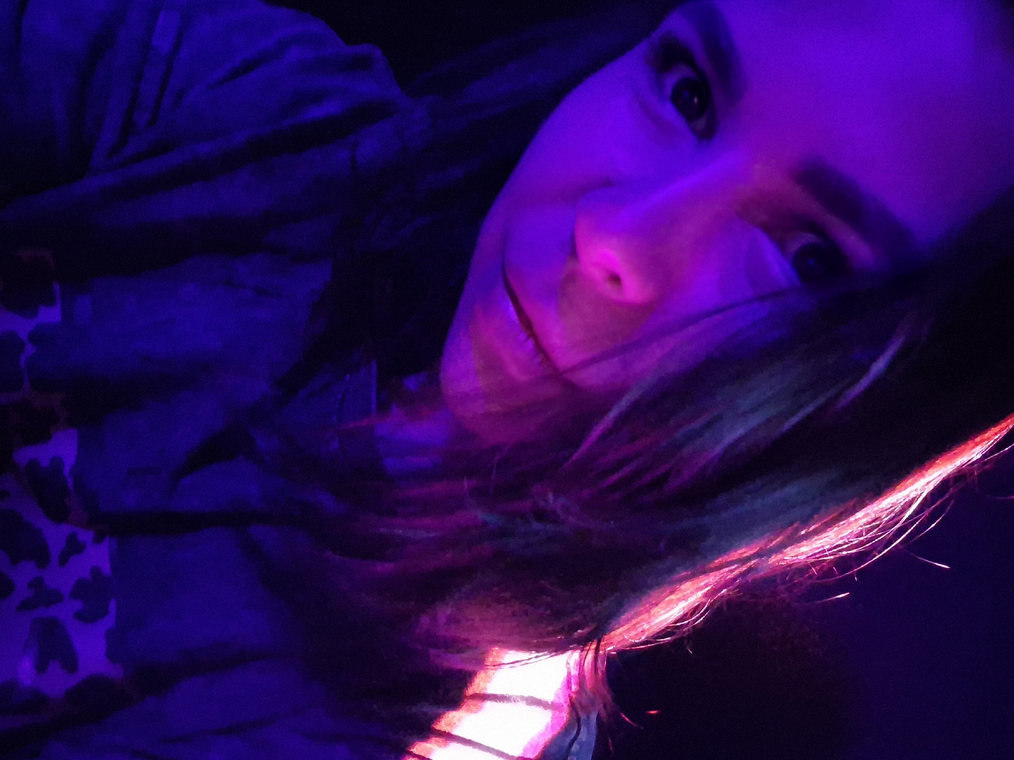 An eye-level, tilted close-up photograph captures a person, likely a young adult female, in a dimly lit indoor setting, possibly in Tobolsk, Russia. The individual is positioned diagonally across the frame, looking directly at the viewer with a subtle smile. Their long, dark hair is prominent, with strands on the right side intensely illuminated by a vibrant, hot pink or magenta light, creating striking highlights that suggest either dyed hair or strong light reflection.
The person's face is bathed in a predominant purple and pink light, giving their skin a luminous, almost ethereal glow, particularly on their nose and forehead. Their dark eyes are open and engaging. They appear to be wearing dark clothing, possibly a hoodie or jacket, and a small section of white fabric with dark, irregular spots (resembling cow print) is visible on the lower left, potentially part of their attire or an item nearby. The background is dark and indistinct, emphasizing the subject and the dramatic, artificial lighting. The scene suggests an evening or night setting with special lighting, such as from neon signs or a club environment. No text is visible in the image.