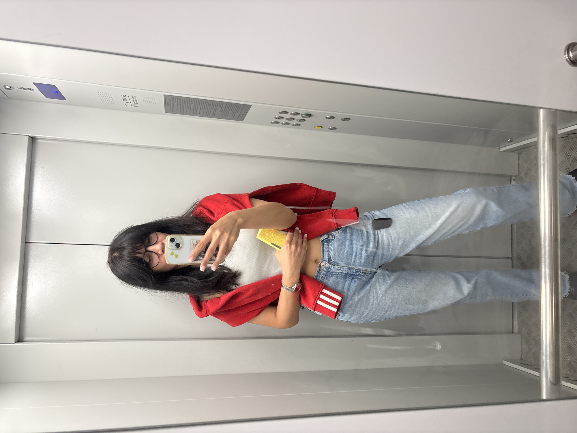 A young woman stands inside an elevator, likely taking a selfie in a mirror. She wears a red hooded sweatshirt, a white t-shirt, and light blue jeans. The elevator interior is modern, with gray walls, a metal control panel with buttons, and a textured floor. The location is impossible to determine based on this single image.