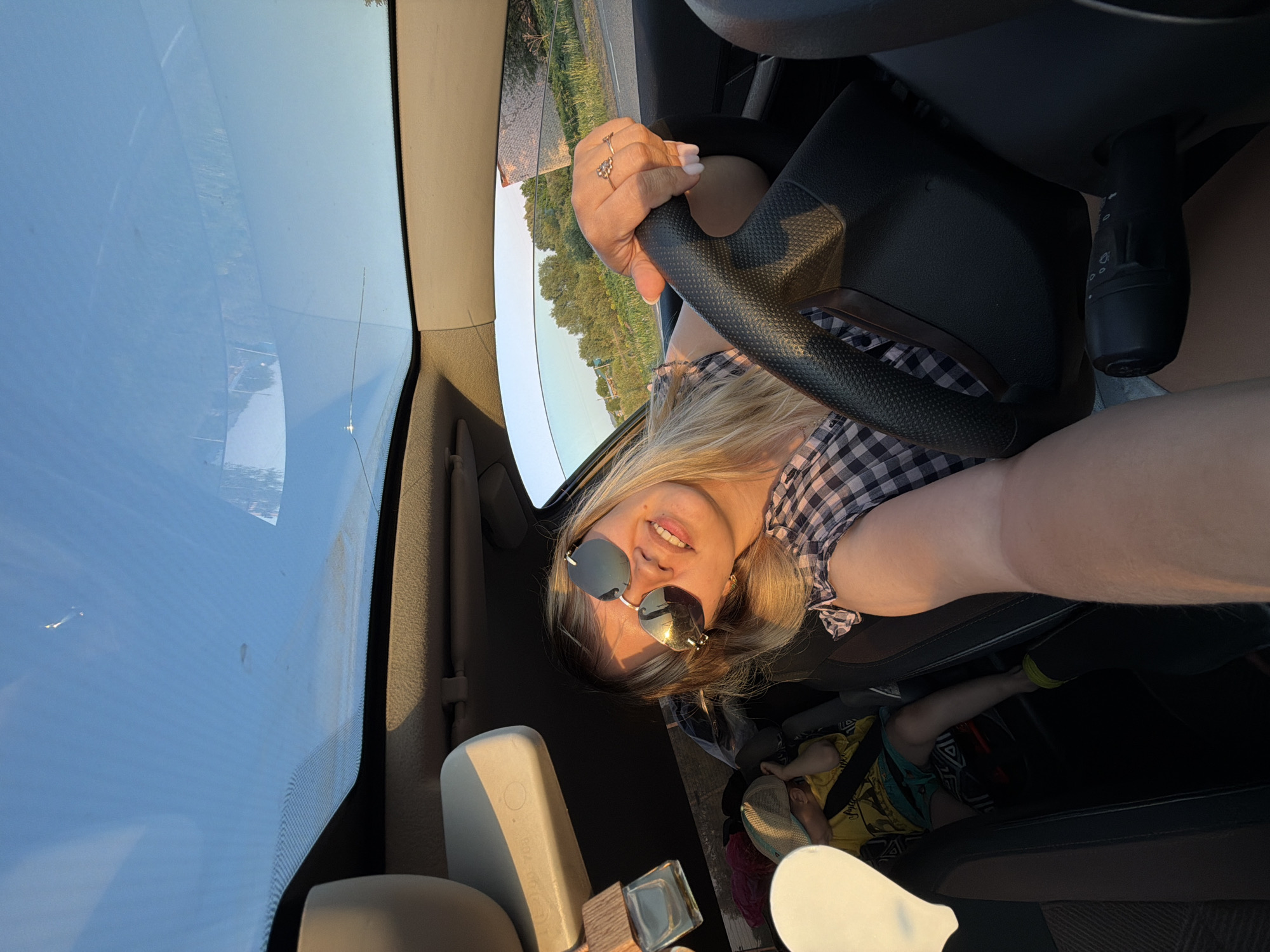 A woman is sitting in the driver's seat of a car, holding the steering wheel with her left hand and taking a selfie. She is wearing sunglasses and a checkered shirt. Through the window, trees and sky are visible. In the backseat, a child lies down. The location is not specified as Чумна, Russia.
