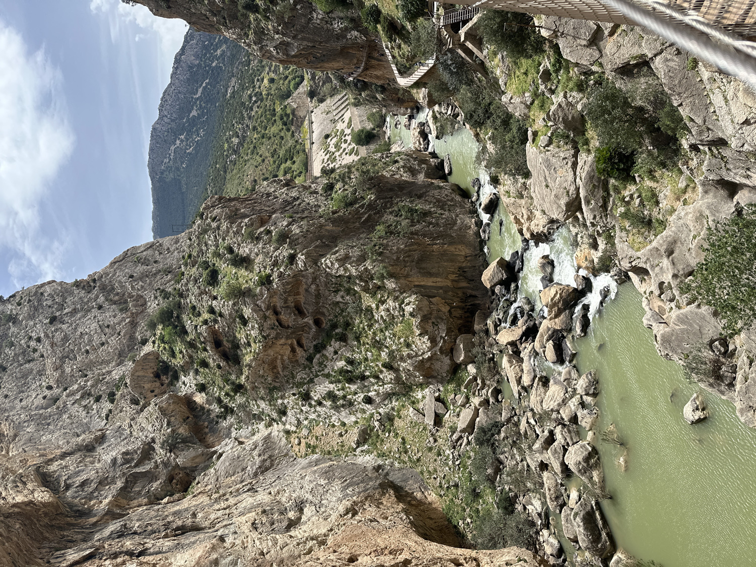 This media file presents a panoramic view of a deep, rugged river gorge, characteristic of the Caminito del Rey near Ardales, Spain. Towering, stratified rock cliffs dominate the scene, descending sharply to a river of greenish-turquoise water flowing at the bottom. The river's course is dynamic, featuring sections of white-water rapids where it tumbles over large boulders, interspersed with calmer, reflective pools.
A prominent feature is the narrow, suspended walkway, likely part of the Caminito del Rey, which is visible clinging to the upper right cliff face and extending into the distance on the left. This pathway is constructed from wooden planks supported by metal structures and cables, offering a dizzying perspective into the canyon. Throughout the cliffs, particularly on the lower left, numerous small, dark openings or 'holes' can be observed in the rock formations, hinting at natural caves or geological features.
Sparse but resilient green vegetation, including shrubs and small trees, sprouts from crevices on the cliff faces and lines parts of the riverbank, contrasting with the predominantly grey and brown rock. The sky above is bright with scattered white clouds, suggesting a sunny day, likely late morning or early afternoon, as indicated by the clear illumination and visible shadows. Distant mountain peaks rise in the background. The scene evokes a sense of awe and adventure, capturing the scale and dramatic beauty of this natural and engineered landscape. No people or visible text are present.