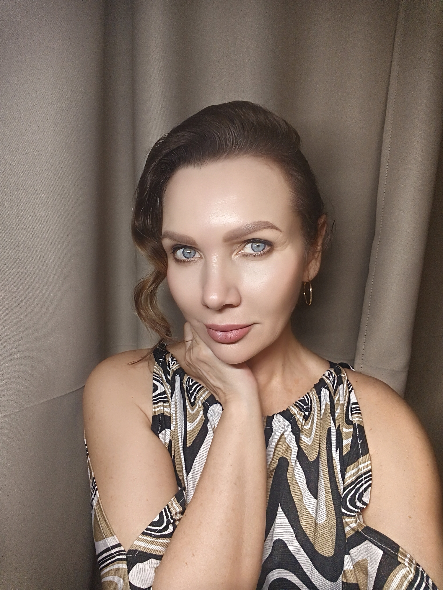 A woman is positioned indoors, her face close to the camera, her right hand gently supporting her chin. She wears gold hoop earrings and a sleeveless top with a geometric pattern of black, white, and gold. Her hair is styled with a side wave. Her light blue eyes are clearly visible. Behind her is a beige backdrop.