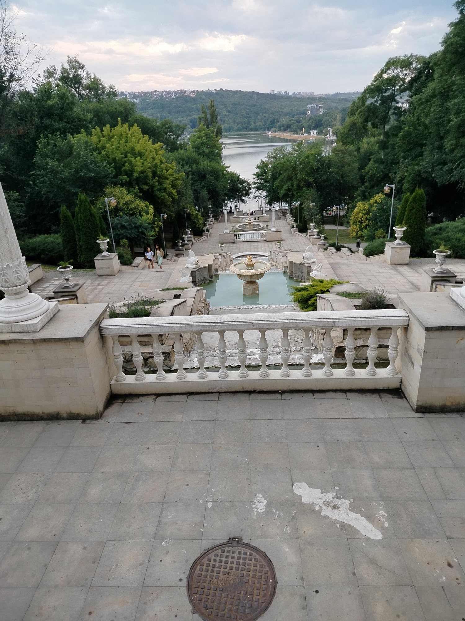 The image depicts a grand staircase leading down towards a lake, surrounded by lush greenery and a city skyline in the distance. This iconic landmark is the Cascades in Chișinău, Moldova.
In the foreground, a wide, tiled staircase stretches downwards, with a weathered manhole cover visible on the bottom landing. A white stone balustrade with ornate pillars lines the sides of the staircase, offering a view of cascading fountains and tranquil pools. The main fountain, centrally located, features a tiered structure with water gently flowing.
Further down the staircase, on a mid-level landing, two young women are seen descending. They are dressed casually, with one carrying a backpack, suggesting a leisurely stroll. The surrounding area is adorned with large decorative urns placed on stone pedestals, interspersed with manicured shrubs and tall, conical evergreens.
Beyond the immediate staircase, the scene opens up to a serene lake, reflecting the overcast sky. The opposite bank of the lake is covered in a dense forest, leading up to a distant cityscape with buildings of various sizes. The overall atmosphere is one of tranquility and natural beauty, integrated with architectural grandeur. The lighting suggests it is daytime, with the sky being cloudy.