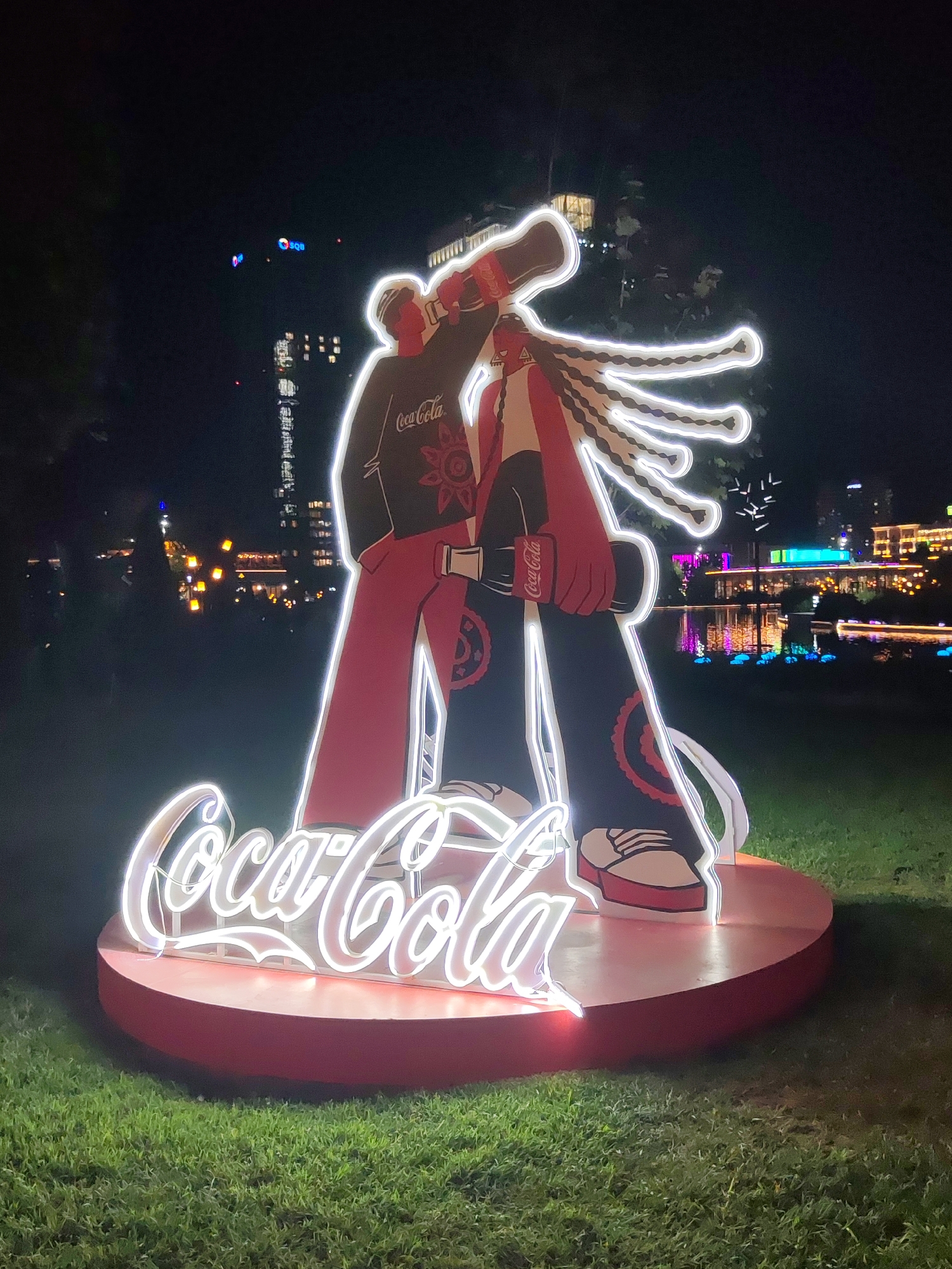 An illuminated Coca-Cola advertisement is displayed on a grassy area at night, in the city of Tashkent, Uzbekistan. The advertisement features two cartoonish figures, one male and one female, dressed in stylized clothing with Coca-Cola branding. The figures are embracing and holding a Coca-Cola bottle between them. The Coca-Cola logo is prominently displayed in neon lights at the base of the advertisement. In the background, city lights and a body of water with reflections are visible. The overall scene is vibrant and festive, likely part of a promotional event or installation.