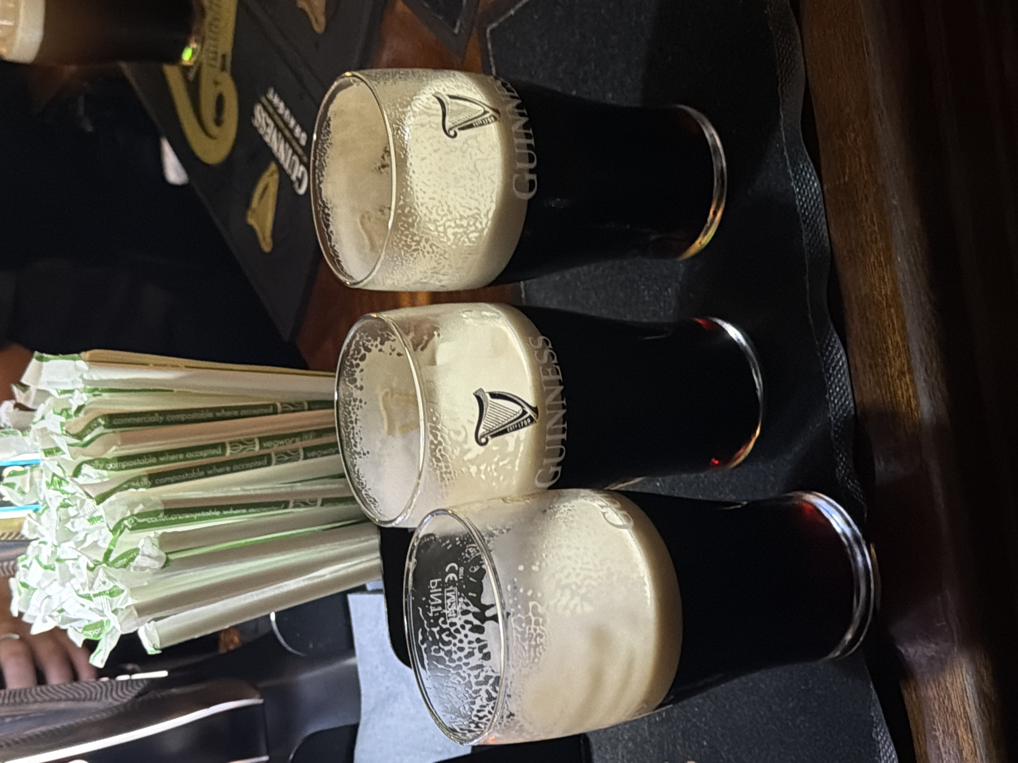 The image shows three glasses of Guinness stout with a thick, creamy head, placed on a black mat, likely at a pub or bar. To the left, there's a stack of wrapped straws, marked as "commercially compostable." The brand "GUINNESS" is visible on the glasses. The setting appears to be indoors, possibly in London, United Kingdom, though the precise location cannot be identified based solely on the provided image. No people are clearly depicted.