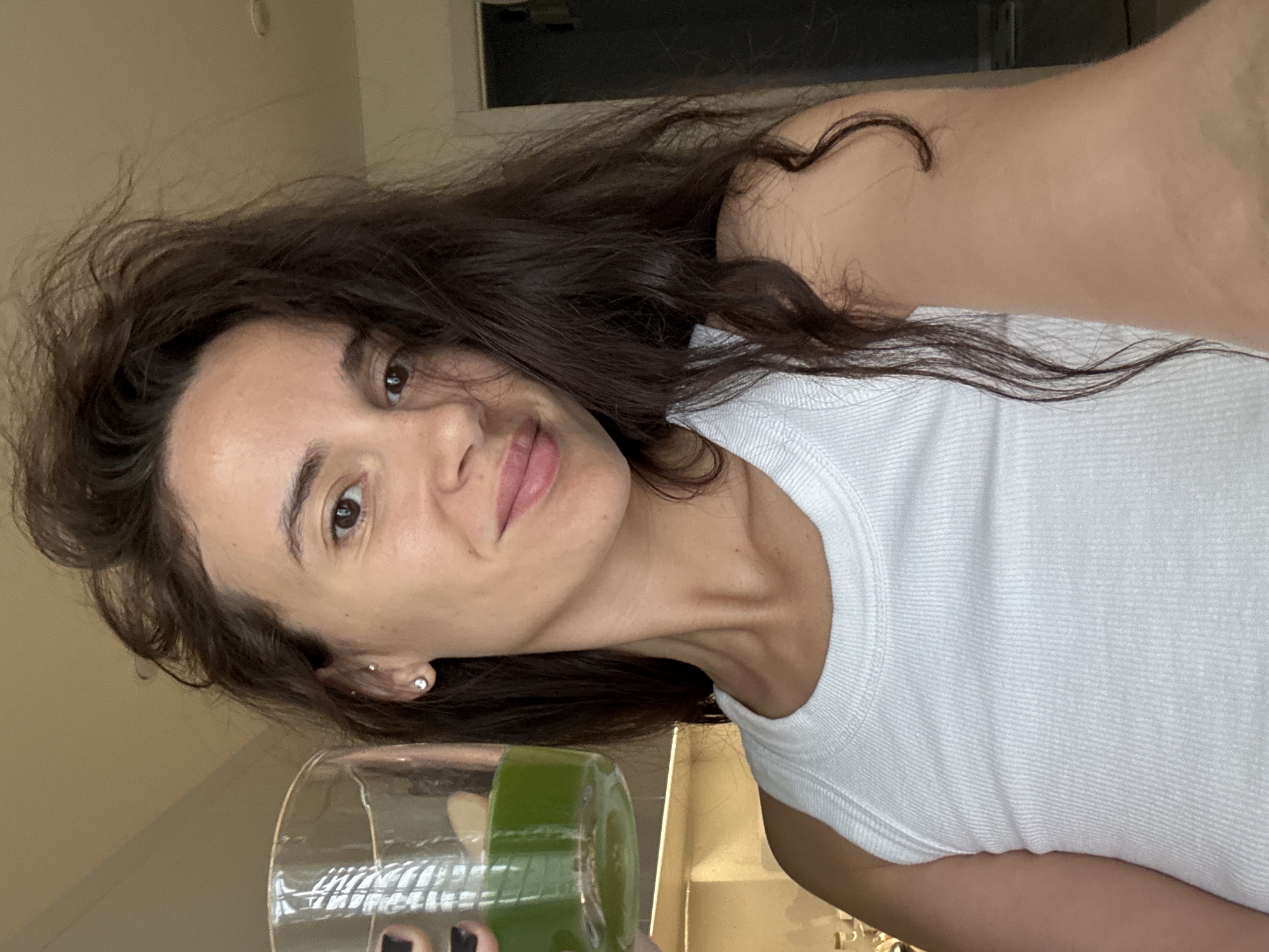A woman with long, dark brown, tousled hair is taking a selfie indoors. She has a natural appearance, featuring a slight smile and visible small stud earrings in her left ear. She is wearing a white ribbed tank top. In her left hand, which has dark-painted fingernails, she holds a clear glass filled approximately halfway with a vibrant green liquid. The background is an indistinct, light-colored indoor space, likely a residence, with reflections visible on the glass suggesting the presence of windows or a strong light source. No specific geographical location within Chaikovsky, Russia, or any other region, can be identified from the visual information provided.