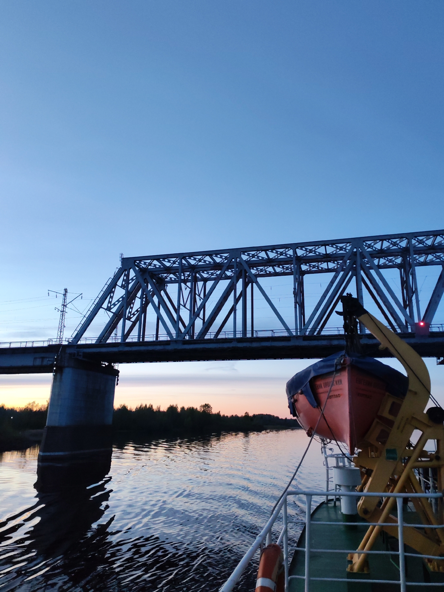 The image shows a view from a boat on a river, with a large steel truss bridge spanning the waterway. The bridge has a concrete pier on the left side of the frame and an electrical pole on the deck, and appears to be for rail use. An orange lifeboat, partly covered with a blue tarp, hangs from a davit on the deck of the boat. The sky is clear with a gradient from light blue near the top of the frame to orange near the horizon. Trees line the shore in the background. The location is likely Нифантово, Russia, considering the style of the bridge and the presence of Russian text on the lifeboat.