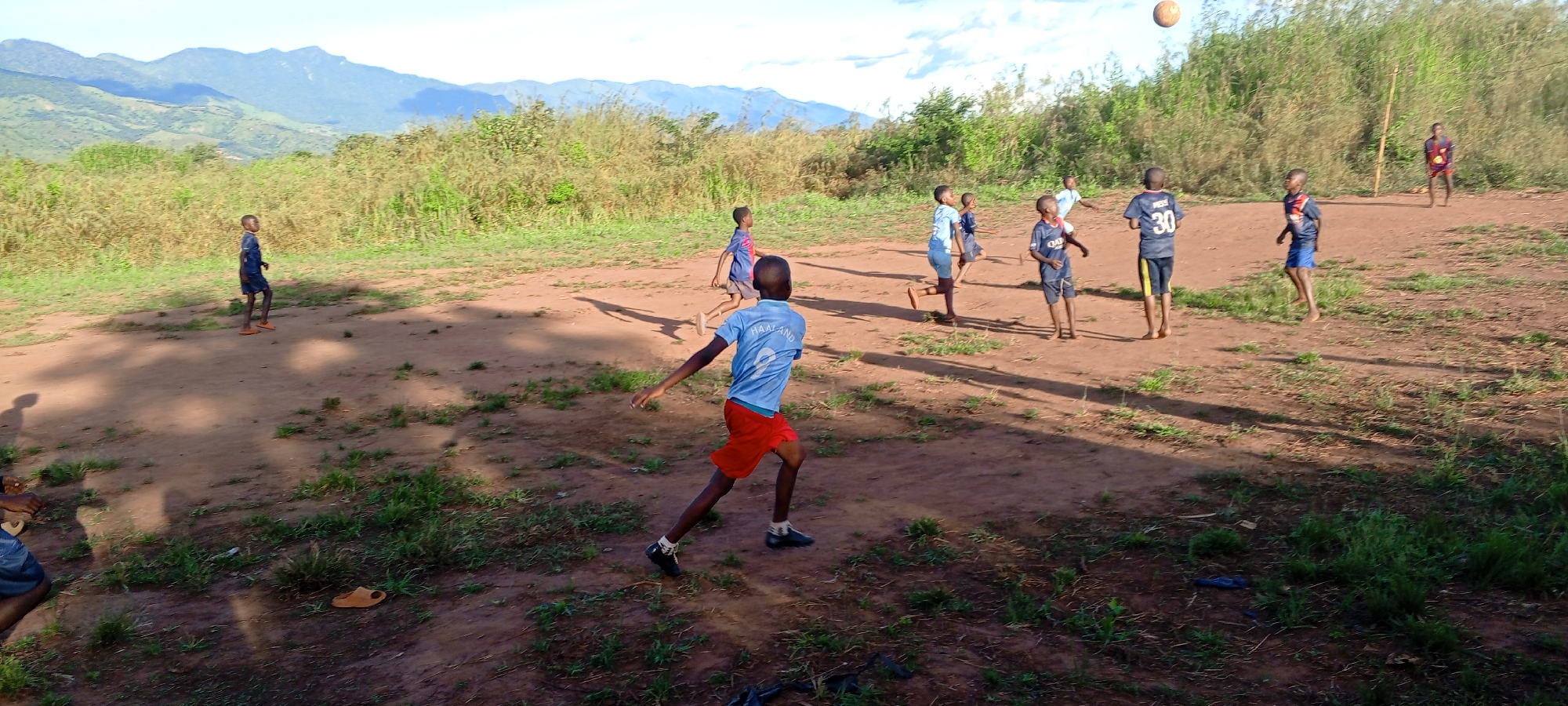 A group of boys plays soccer on a dirt field in Ngalula, Democratic Republic of the Congo. The player in the foreground is wearing a light blue shirt with the name "Haaland" and the number "9" on the back, along with red shorts. The ball is in the air above the group of boys in the background, most of whom are barefoot. Mountains are visible in the distance.