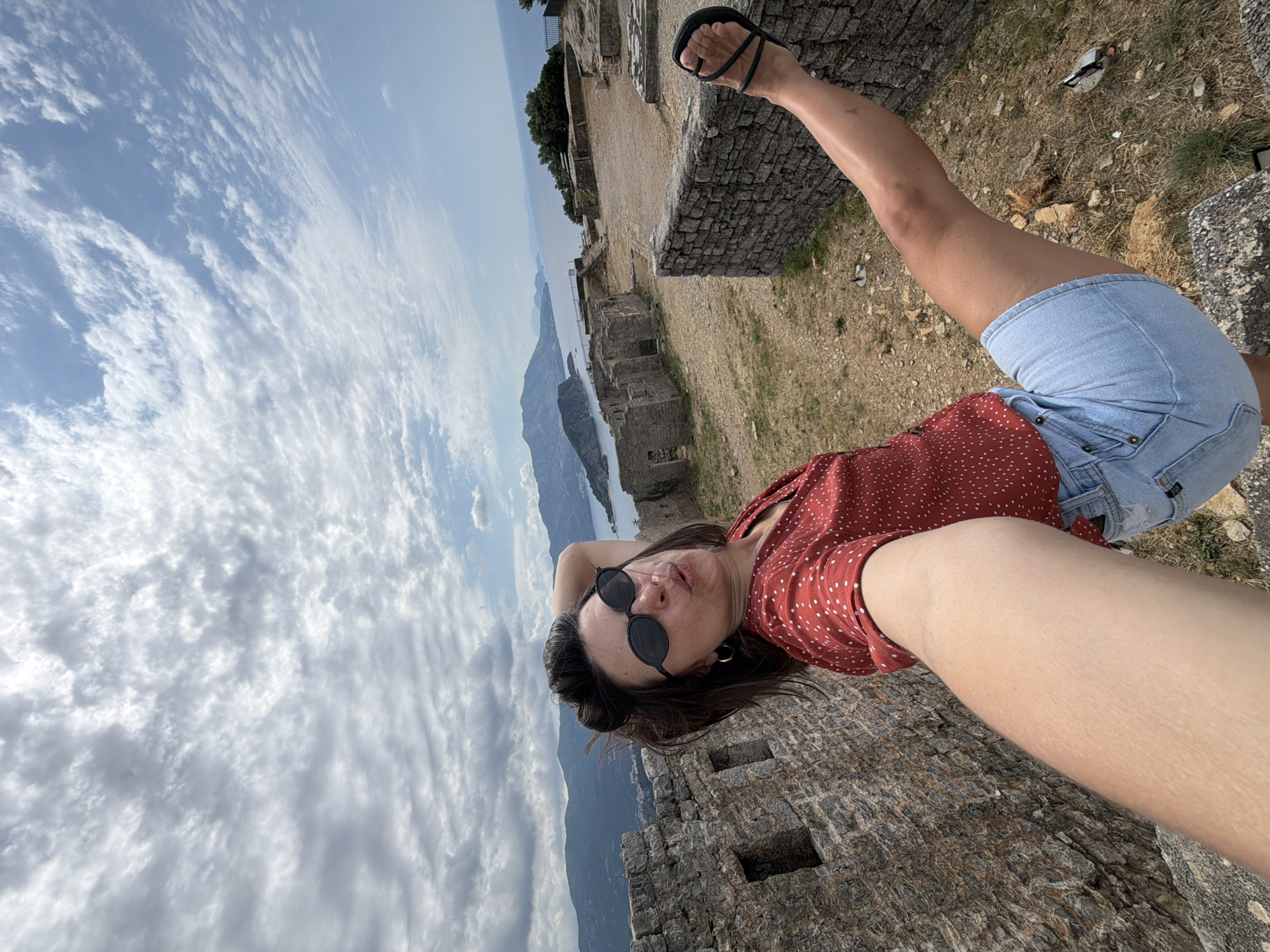The image features a woman posing for a picture, likely a selfie, outdoors on what appears to be ancient ruins. She is positioned diagonally in the frame, lying on her back with her head towards the top left and her feet towards the bottom right. She wears a red top with white polka dots, light blue denim shorts, and black sandals. Dark, round sunglasses rest on her face, and her expression is neutral. Her right leg is bent and raised, with her foot visible in a sandal.
The scene is set at a historic location overlooking a body of water and mountains, characteristic of a coastal area like Budva, Montenegro. Directly behind and below the woman are weathered stone structures and dry, earthy ground. In the background, a vast expanse of water, possibly a bay or the sea, meets a distant mountainous coastline. The sky above is largely covered with white and gray clouds, suggesting an overcast but bright daytime. No discernible text is visible in the image.