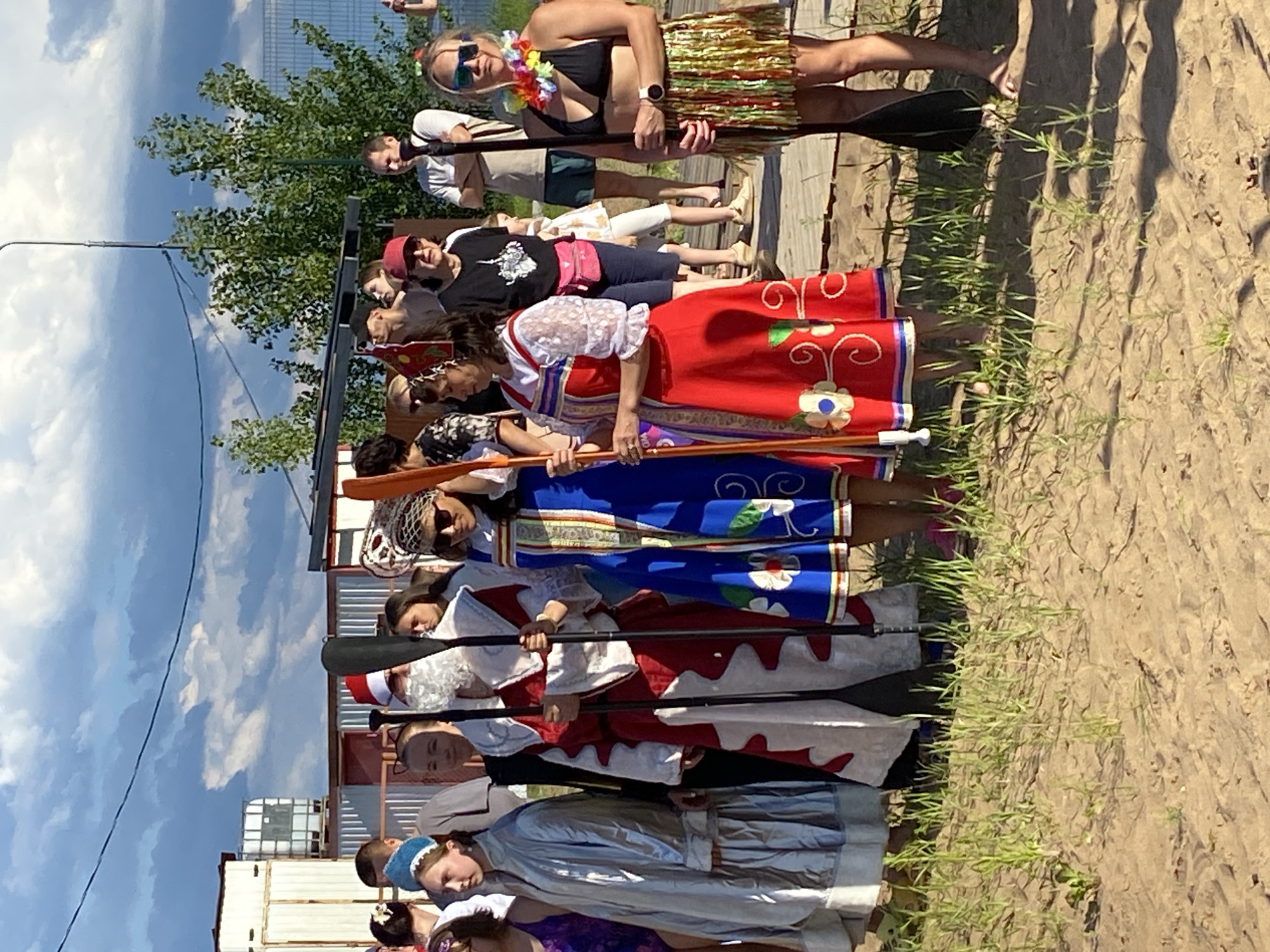 A group of people, some in costume, stand on a sandy embankment near a body of water. The person on the far left is dressed in a silver, hooded robe. Next to them, a person in a red and white Santa Claus costume holds two paddles. To their right, a woman wears a blue and white traditional Russian dress, holding a paddle. Further right, another woman is dressed in a red traditional Russian dress with white lace sleeves, also holding a paddle. The person on the far right is wearing a black bikini top, a colorful lei, and a fringed skirt, holding a paddle. The background shows a cloudy sky, trees, and some industrial-looking structures, possibly part of a port or marina in Chaikovsky, Russia. The sand is covered with sparse green grass.
