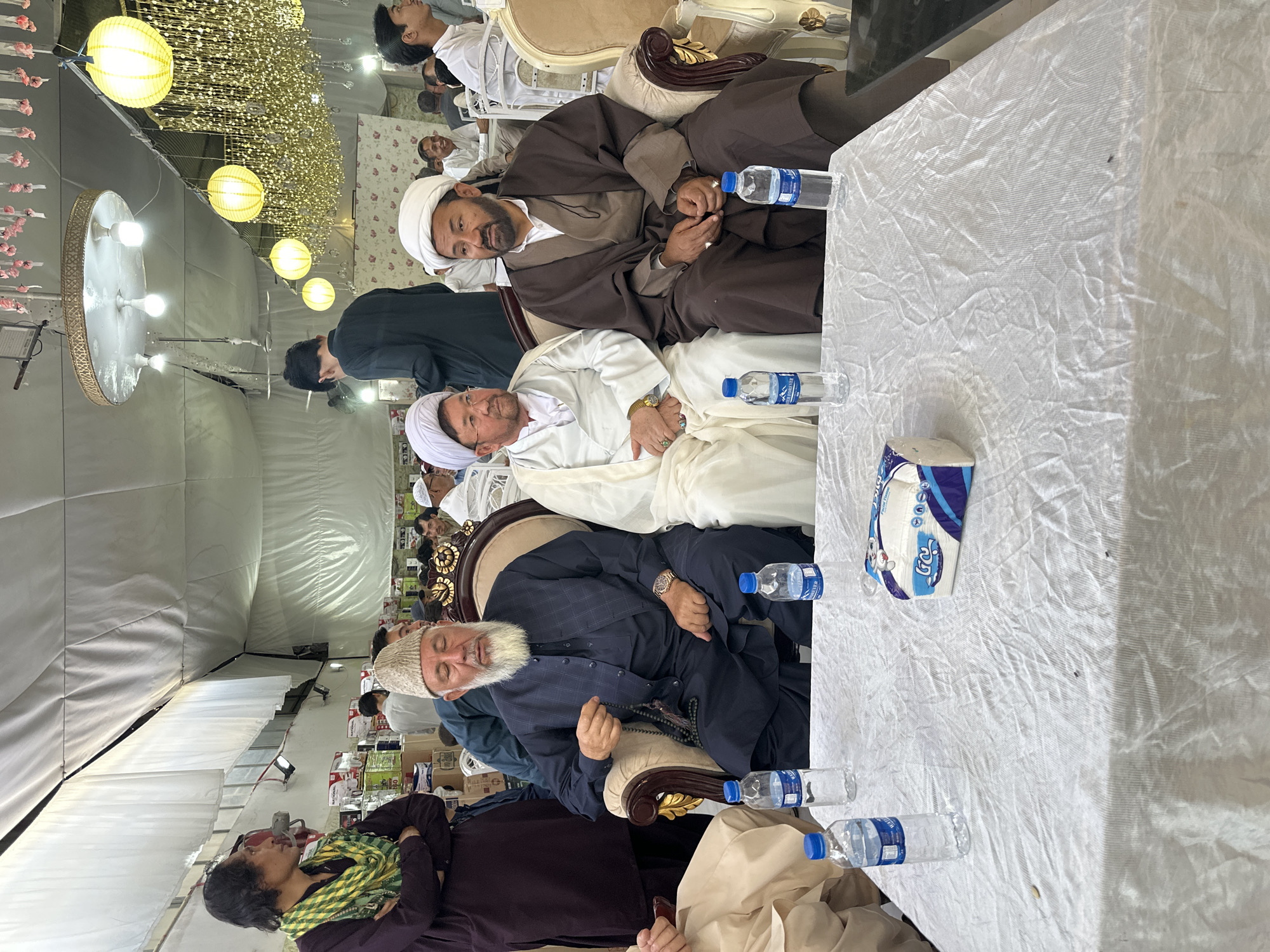 The image shows a group of men sitting at a table in what appears to be a large indoor event space, possibly a tent or hall. The space is decorated with chandeliers and string lights.
The men are dressed in various traditional attire, including robes and turbans, indicating they may be religious figures or community leaders. Several bottles of water are placed on the table, along with a box of tissues. The table is covered with a white cloth.
The presence of these individuals and the setting suggest that this could be a gathering for a religious event, meeting, or celebration. Given the prompt specifying Quetta City Tehsil, Pakistan, it is reasonable to assume that the image was taken in that location.