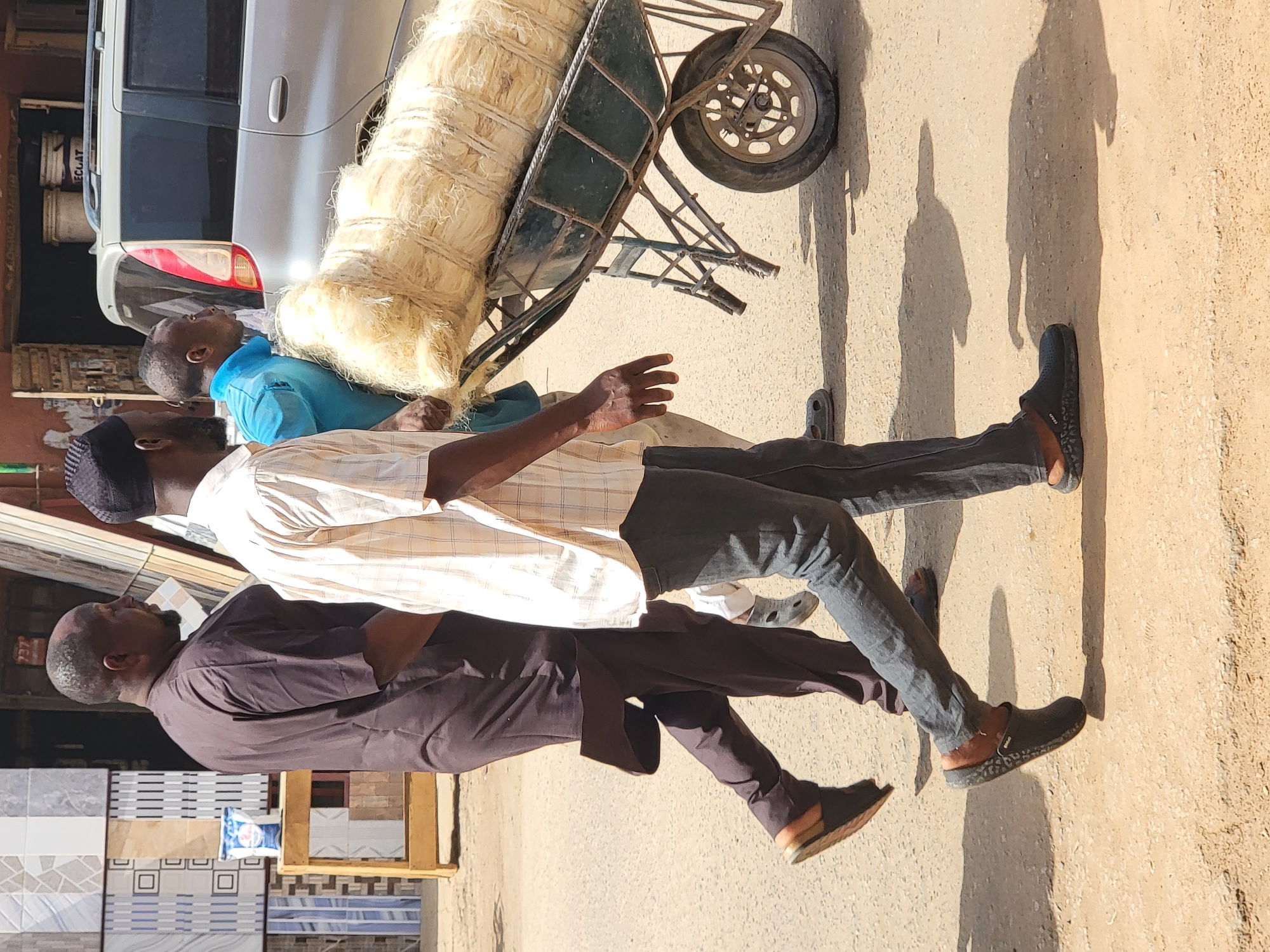 The image depicts a sunny outdoor street scene in Maiburiji, Nigeria, likely during the morning or late afternoon due to the strong, elongated shadows cast on the light-colored paved or dirt ground.
There are three men present in the frame. On the far left, a man with a beard is dressed in a dark brown or purple long-sleeved garment and dark pants, seen from the side facing right. In the center, another man wears a white and light brown plaid shirt, dark pants, and dark slip-on shoes; he is wearing a dark cap and facing left. Partially obscured behind a wheelbarrow, a third man in a light blue collared shirt is looking upwards and towards the right.
A sturdy, dark green or blue metal wheelbarrow, heavily loaded with a large amount of light-colored, fibrous material—possibly sisal or dried grass—dominates the center-right of the image. Its single wheel is visible on the right. Behind the wheelbarrow and the man in the blue shirt, a silver car, possibly an SUV or van, is parked. The word "ECLAT" is faintly visible on its rear.
In the background on the far left, stacks of patterned floor tiles or ceramic sheets, displaying various designs (geometric, striped, plain), suggest a shop or stall selling building materials. A wooden frame and a white plastic bag with some blue, illegible text are also near the tiles. On the sole of one of the dark slip-on shoes worn by the man in the plaid shirt, the brand "CROCS" is visible. The scene captures a casual, everyday street activity, possibly involving the movement or sale of goods.